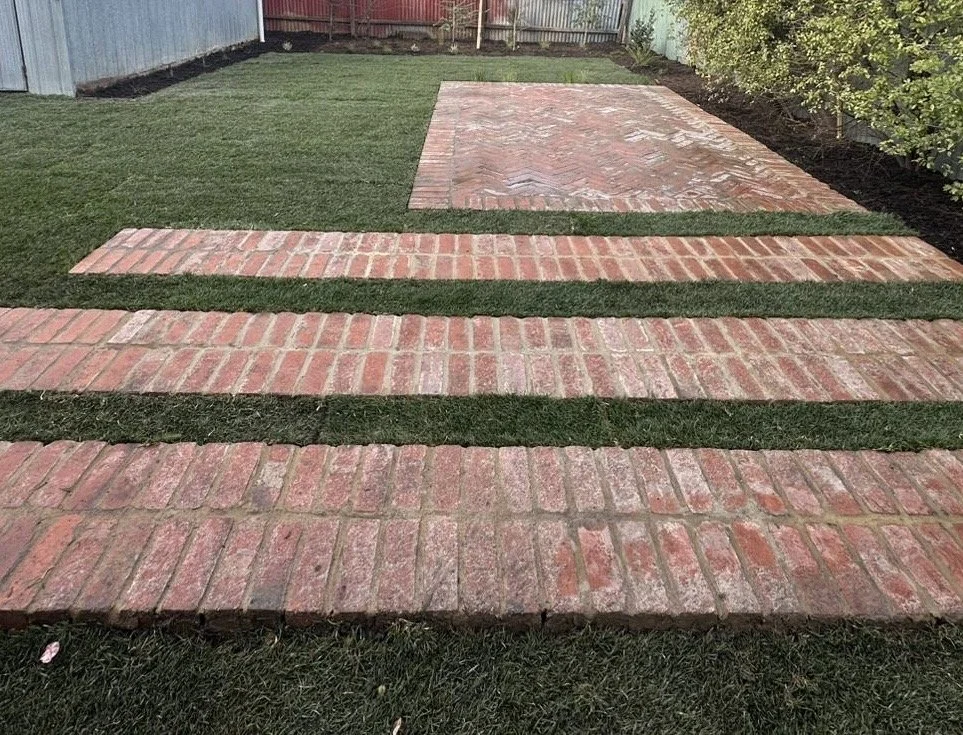 A backyard with a lawn, brick pathway, and brick patio, bordered by a metal fence and some green shrubbery.