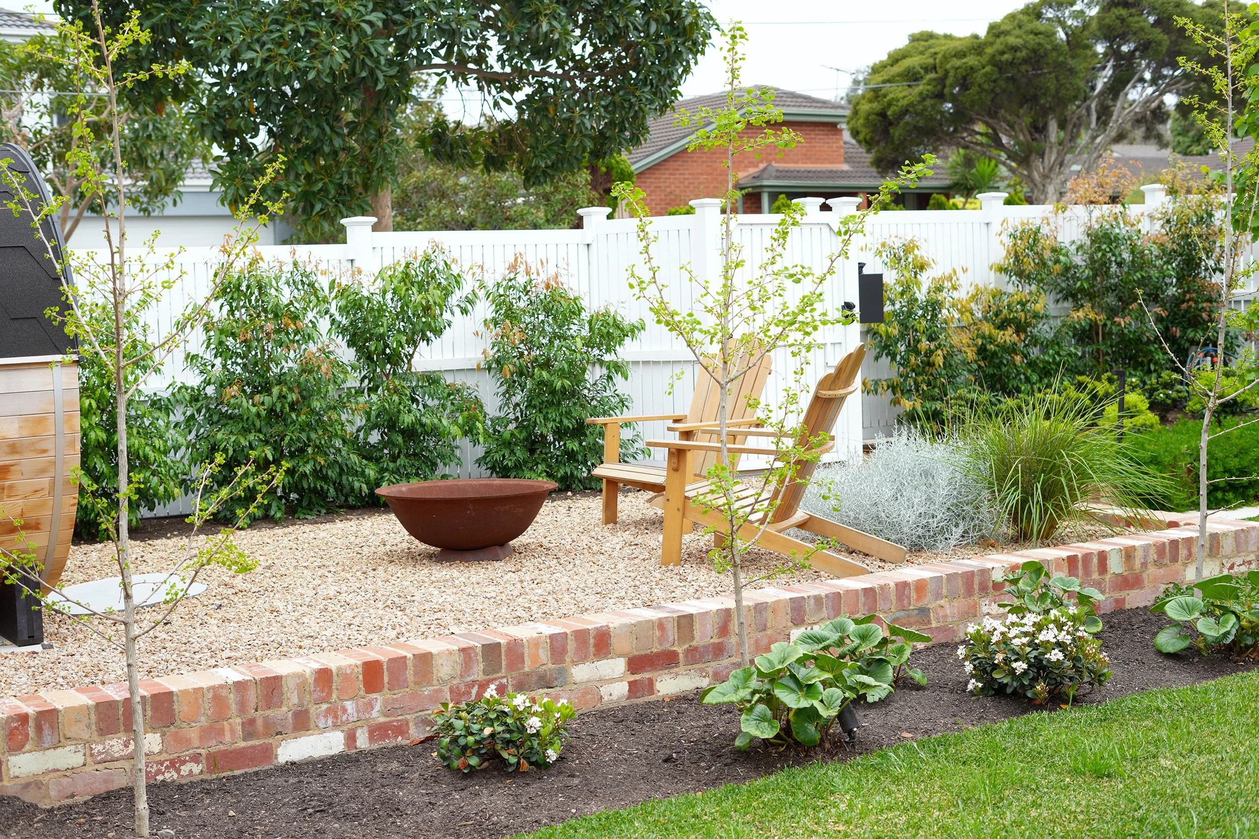 A backyard garden with a white picket fence, various plants and bushes, two wooden chairs, and a small gravel area. There is also a flower bed with blooming plants in front.