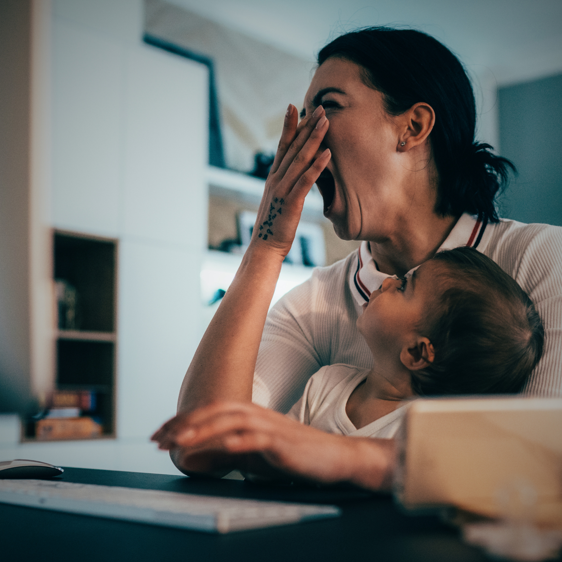 A woman with dark hair yawns while a young child leans on her, looking up at her and smiling, in a home setting.