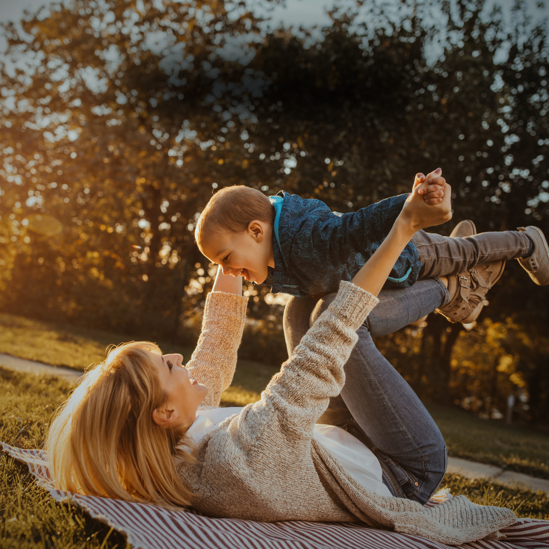 A woman lying on a blanket on the grass playing with her young son outdoors during sunset. The boy is lying on his stomach on her hands, and they are smiling at each other.