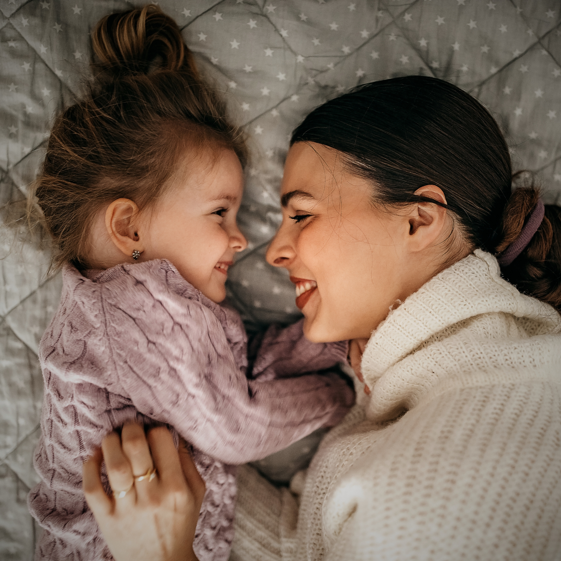 A woman and her young daughter are lying on a bed, face to face, smiling happily at each other.
