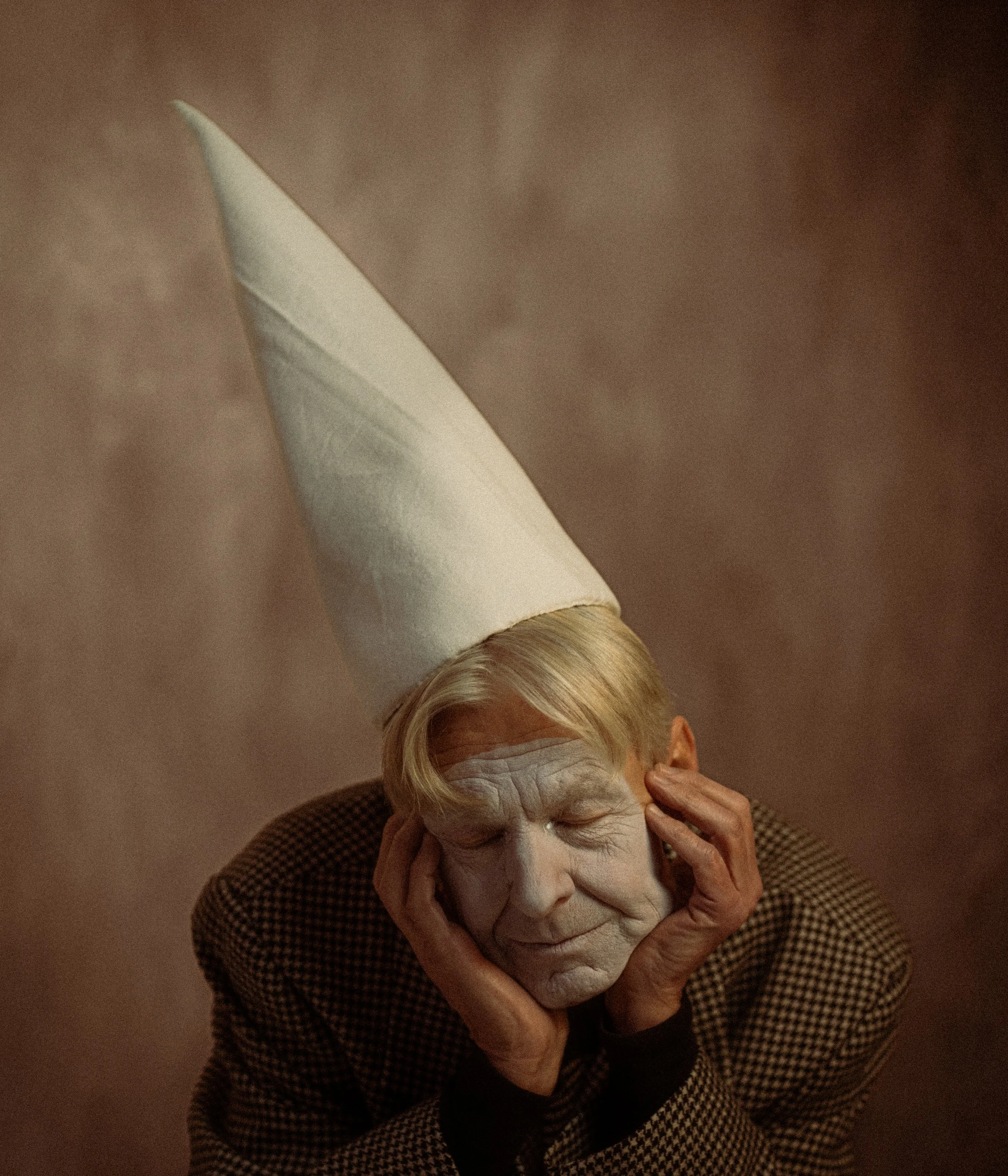 A person with large, prominent facial makeup, white face paint, and blond hair, wears a tall, white conical hat, and has a pensive or sad expression with eyes closed, hands touching the face.