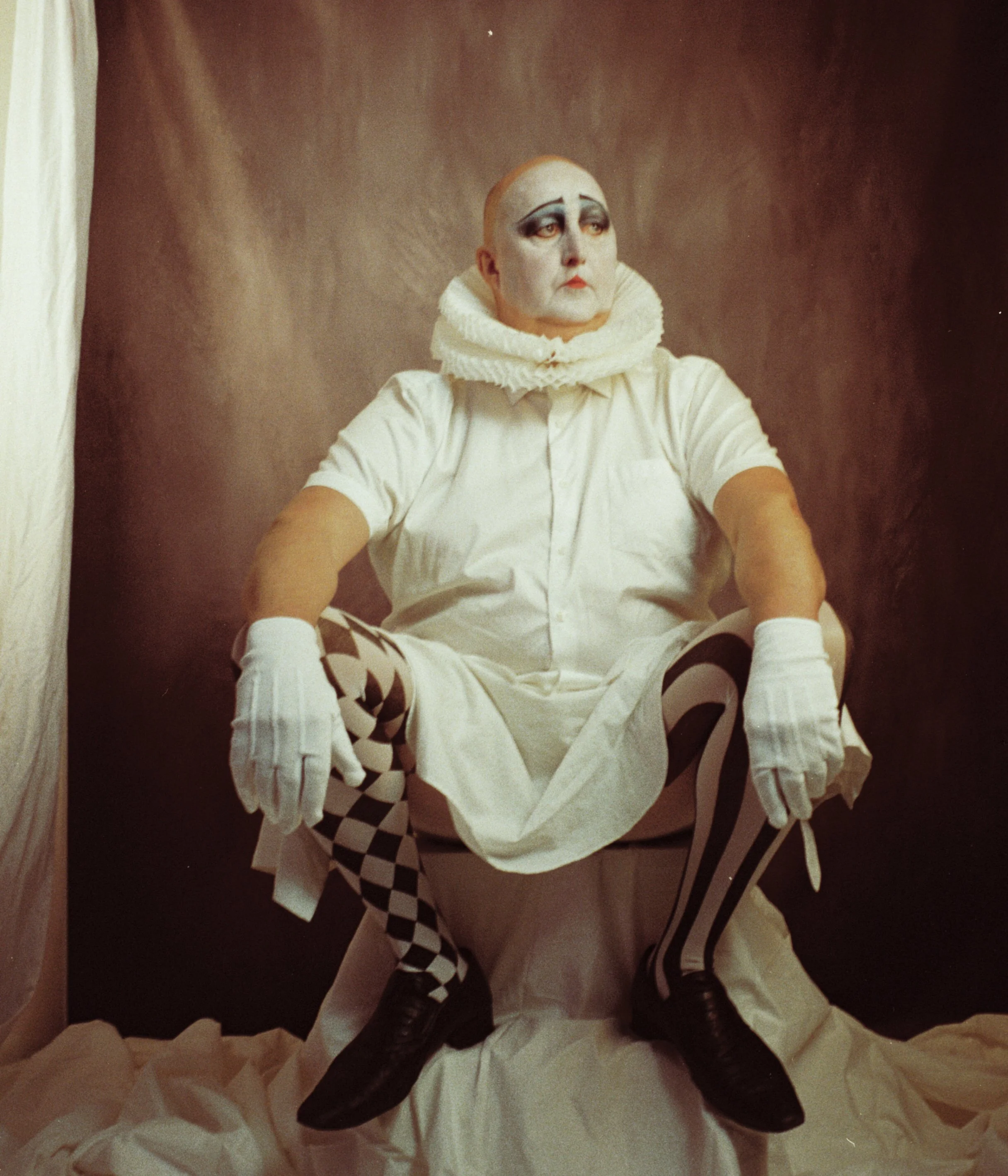 A person dressed as a clown or mime with theatrical makeup, bald head, wearing a white ruffled collar, white shirt, checkered and striped tights, black shoes, and white gloves, sitting on a toilet against a dark background.
