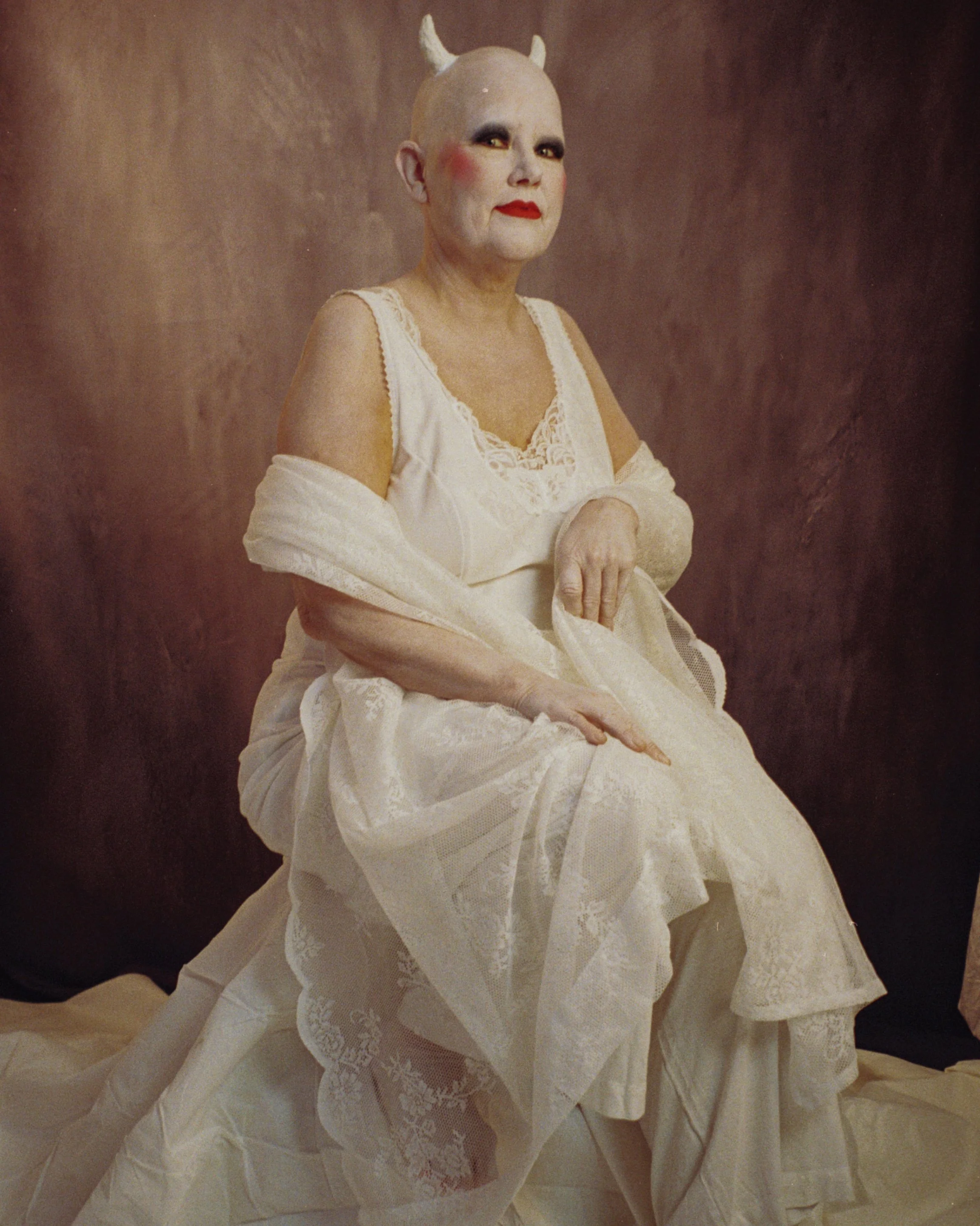 A person sitting on a fabric-covered surface, dressed in a white lace dress, with a pale makeup face, red lipstick, and makeup around the eyes. They have small white horns on their head and are holding part of their dress with one hand. The backgroun