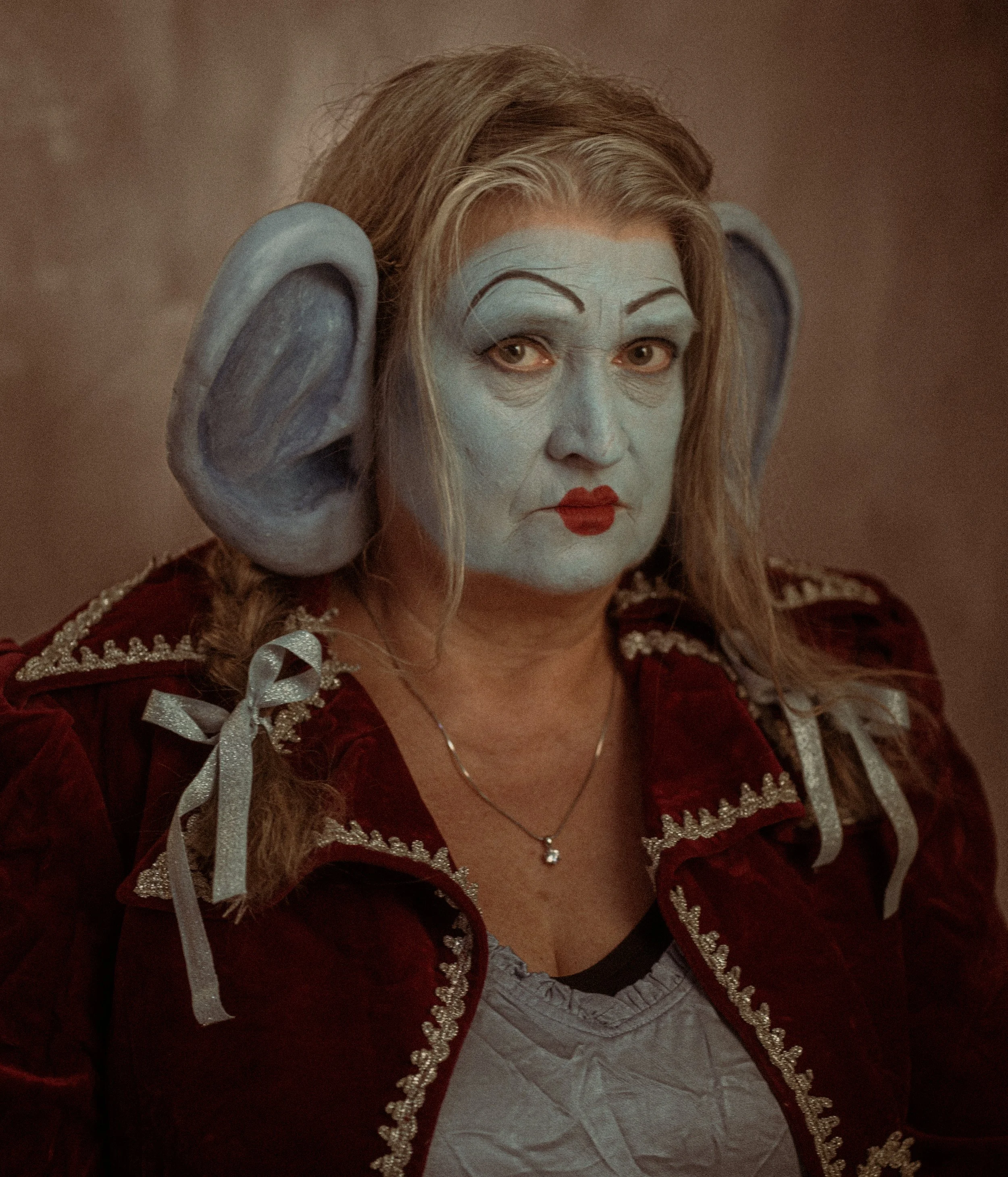 A woman wearing theatrical makeup with blue face paint, exaggerated eyebrows, red lips, and large grey ears costume accessories, dressed in a red velvet jacket with silver accents, and sitting in front of a plain background.