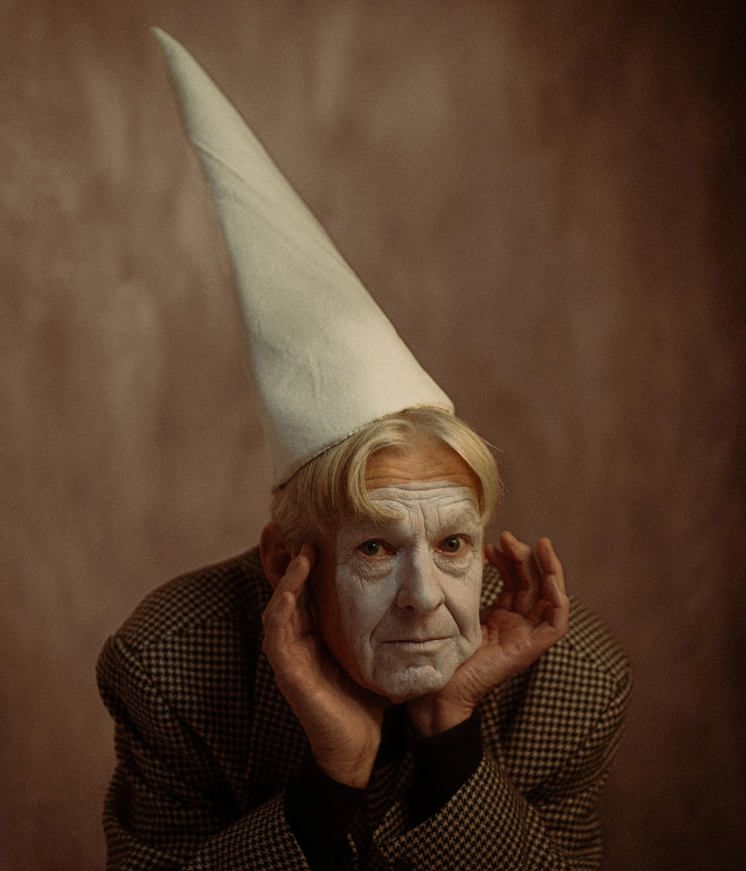 An elderly man with white face makeup, wearing a tall white cone-shaped hat and a checked blazer, rests his head on his hands and looks directly at the camera.