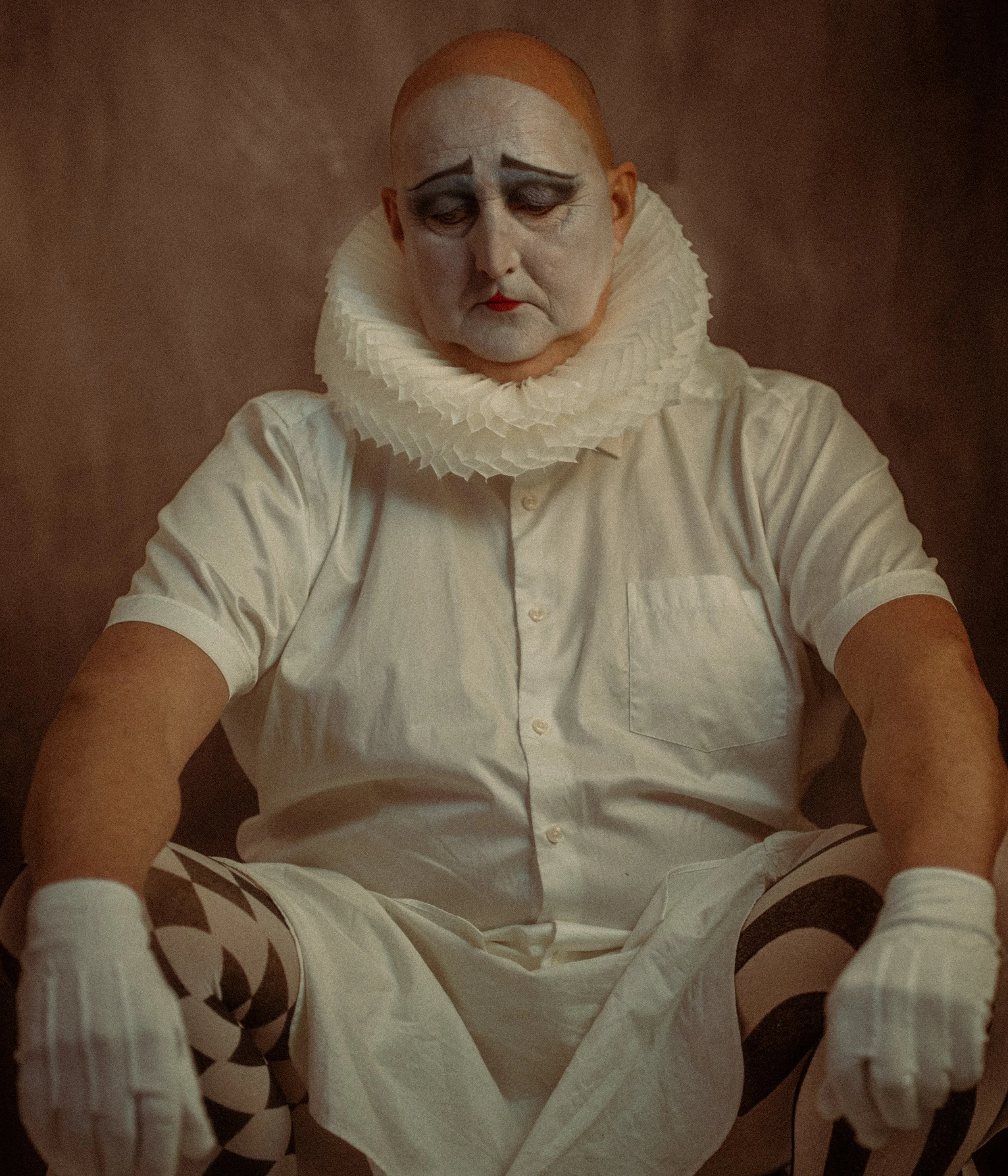 A person dressed as a clown in white with a ruffled collar, makeup with dark eyebrows, and striped tights, sitting with a somber expression.