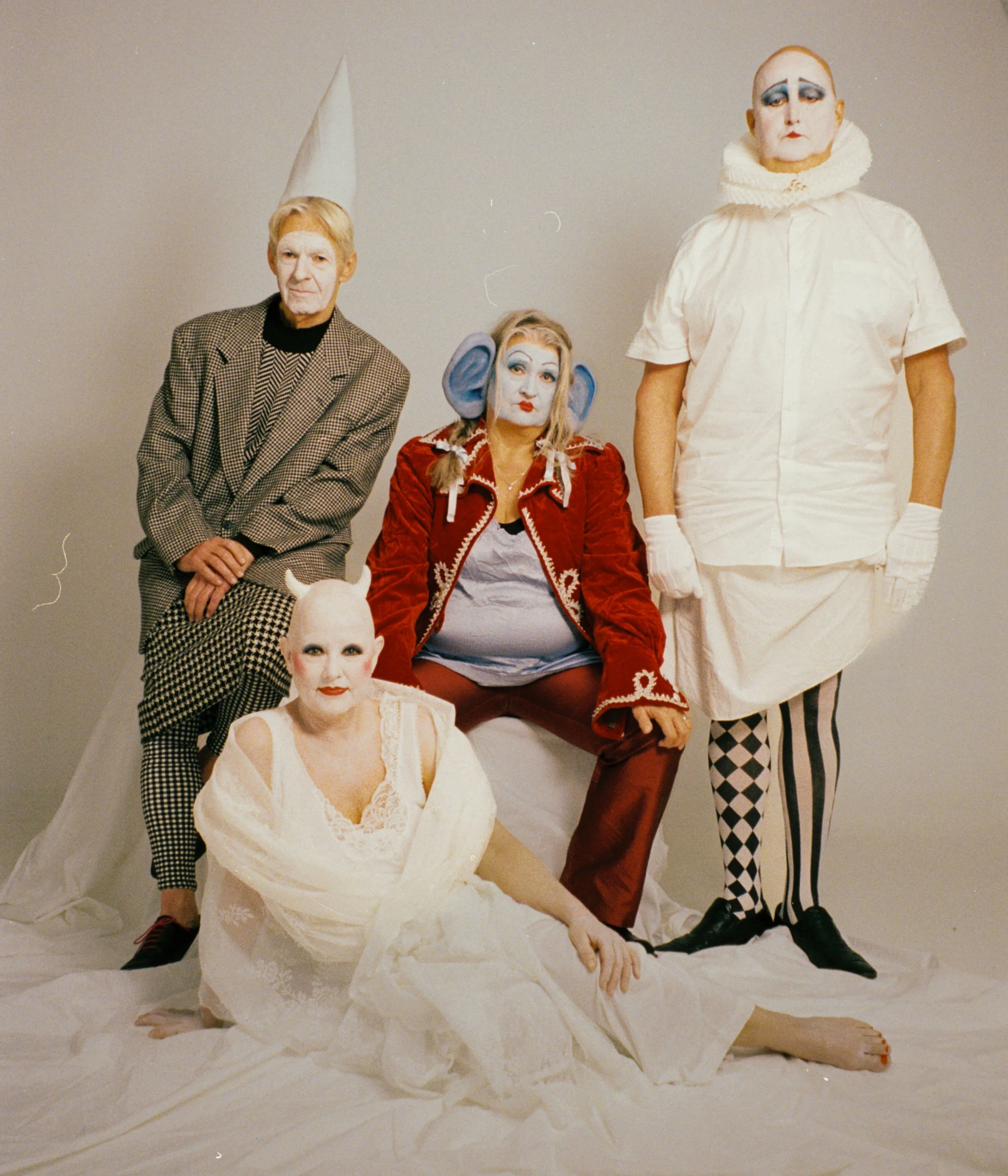 Five performers dressed in clown costumes posing against a plain background. One is seated on the floor, three are standing, and one is seated on a chair. They wear makeup and colorful costumes.