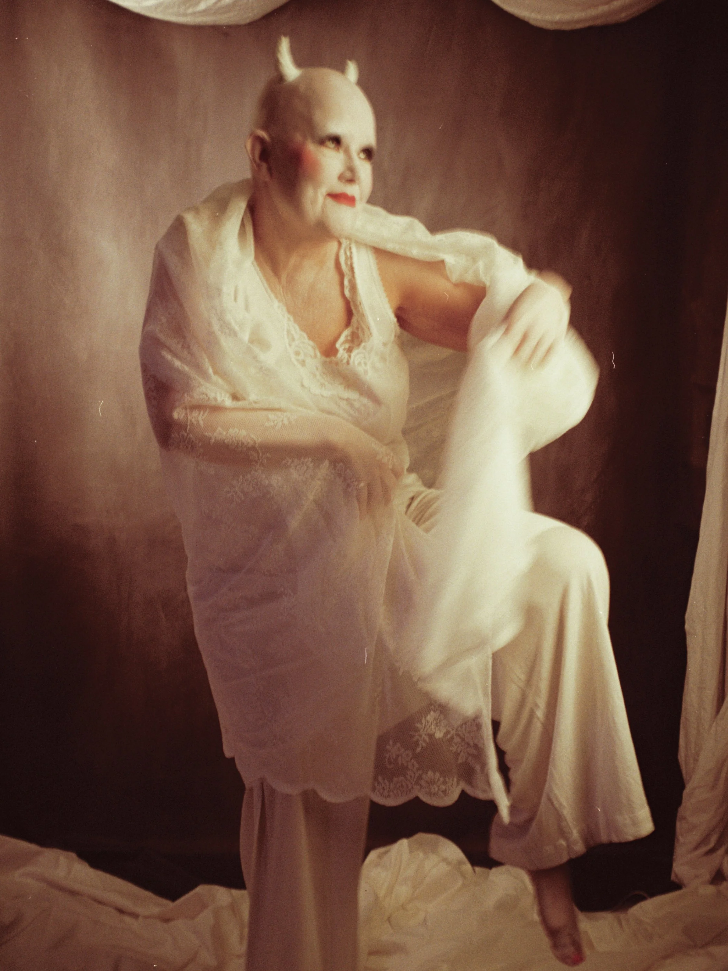 A person dressed in white with lace and sheer fabric, with their hair styled in a bob with small horns, sitting on a stool, raising one leg and resting their foot on a white cloth. The person has makeup with dark eyebrows and red lipstick, and is pos