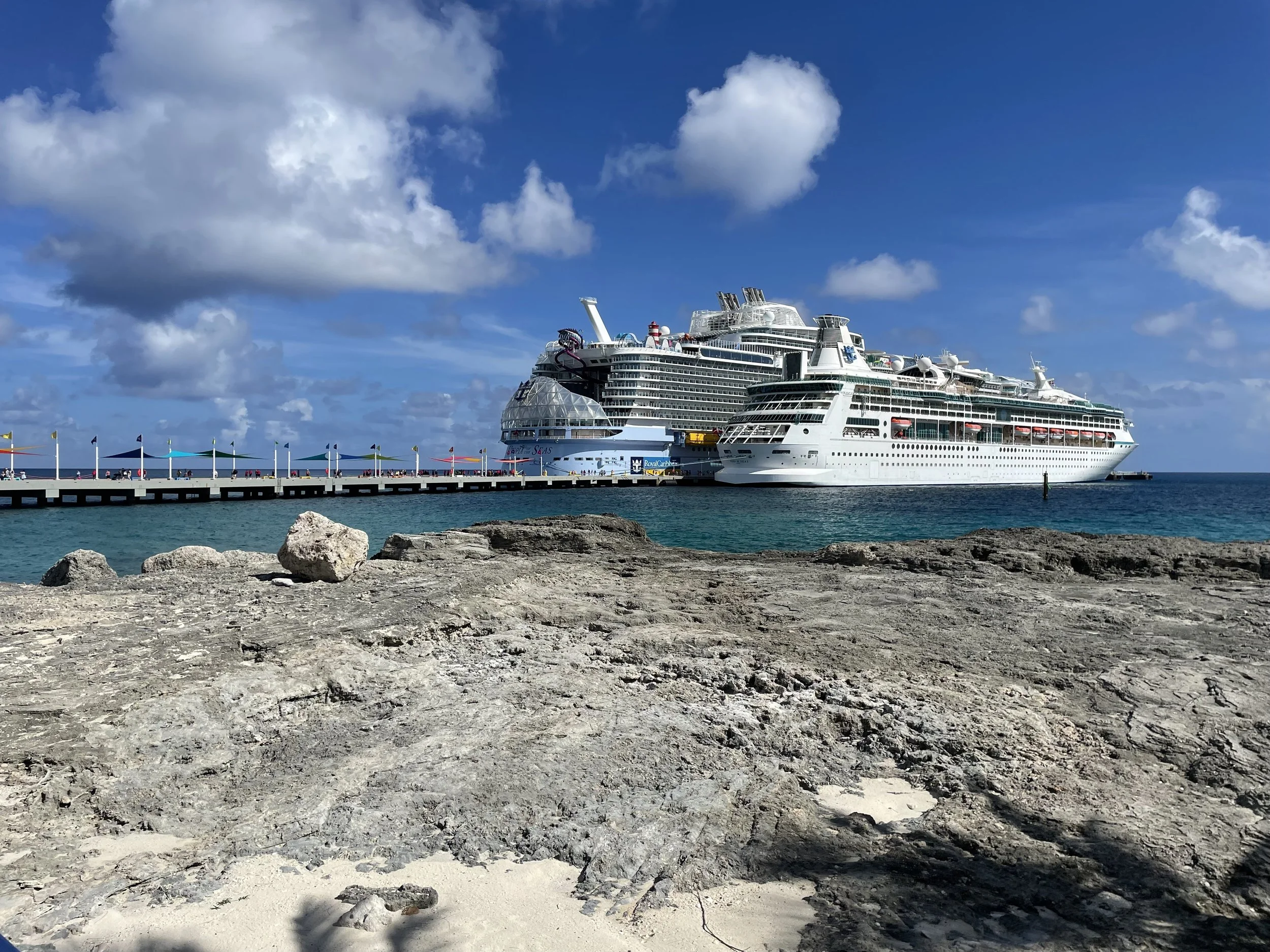 Two cruise ships docked at a pier on a sunny day with blue skies, puffy clouds, calm blue ocean, rocky foreground, and colorful flags along the pier.