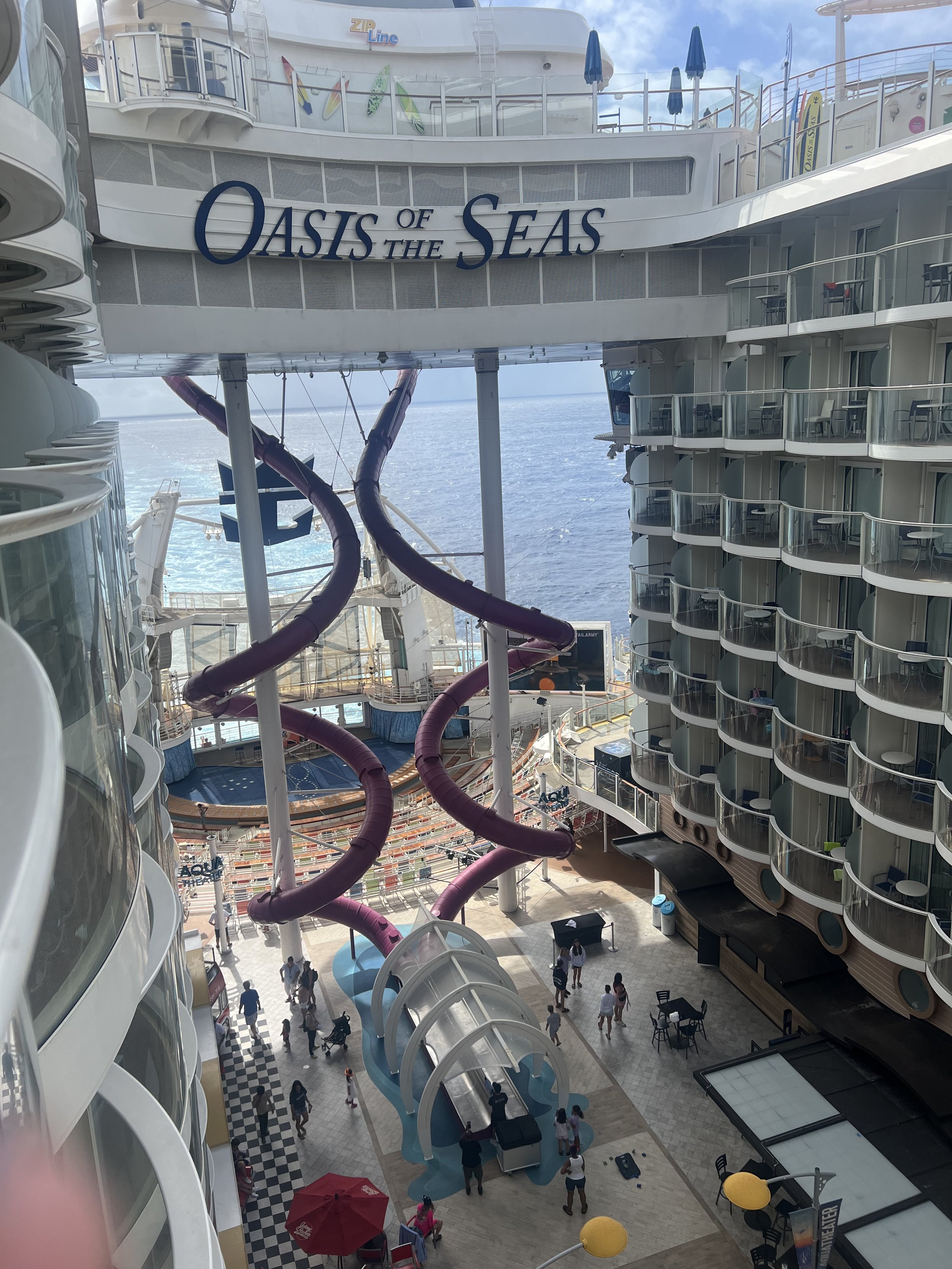View of the Oasis of the Seas cruise ship interior, showing a water slide in the center and the ocean in the background, with multiple balconies and decks.