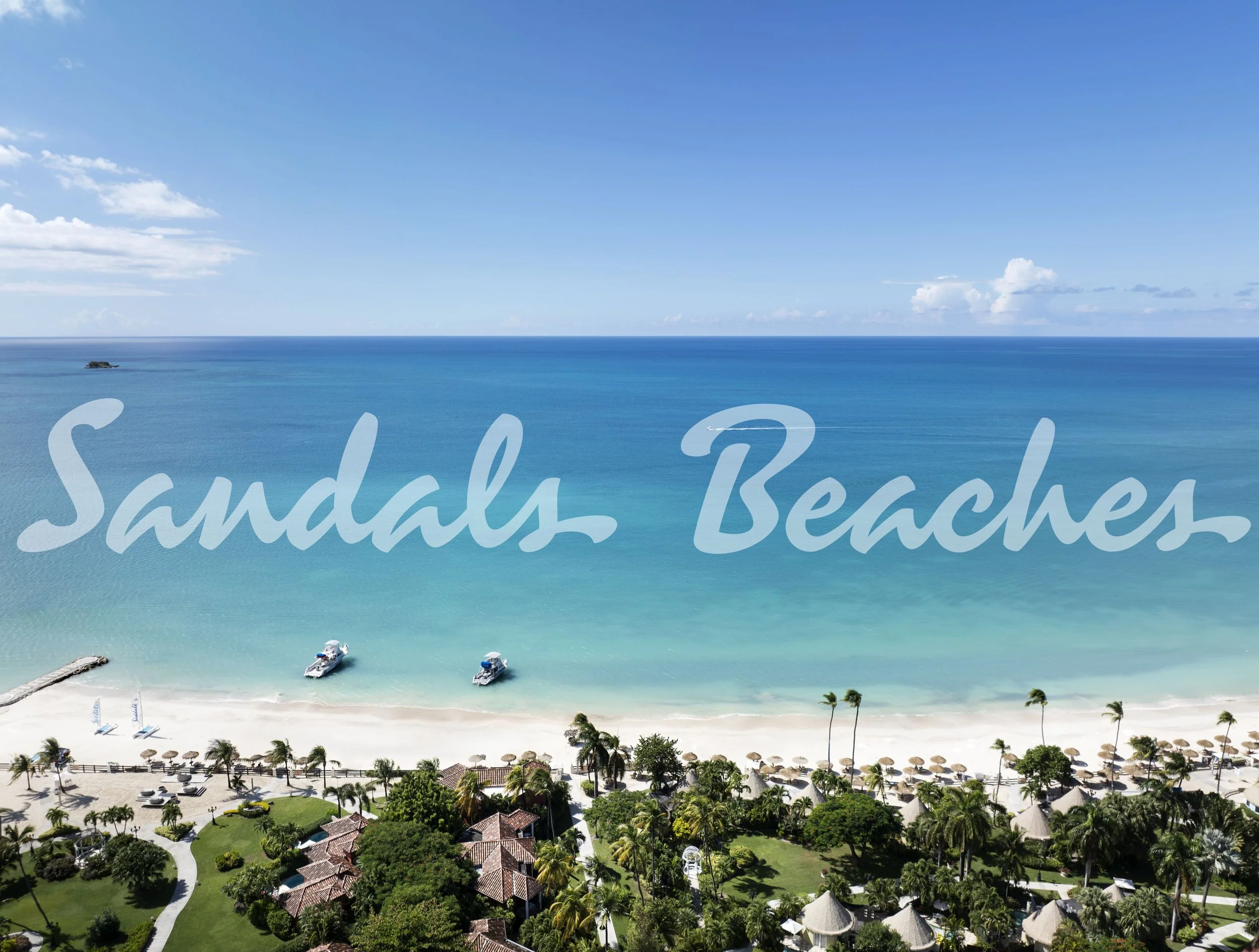 An aerial view of Sandals Beaches Resort with a white sandy beach, turquoise ocean, palm trees, and thatched-roof huts, with a clear blue sky.