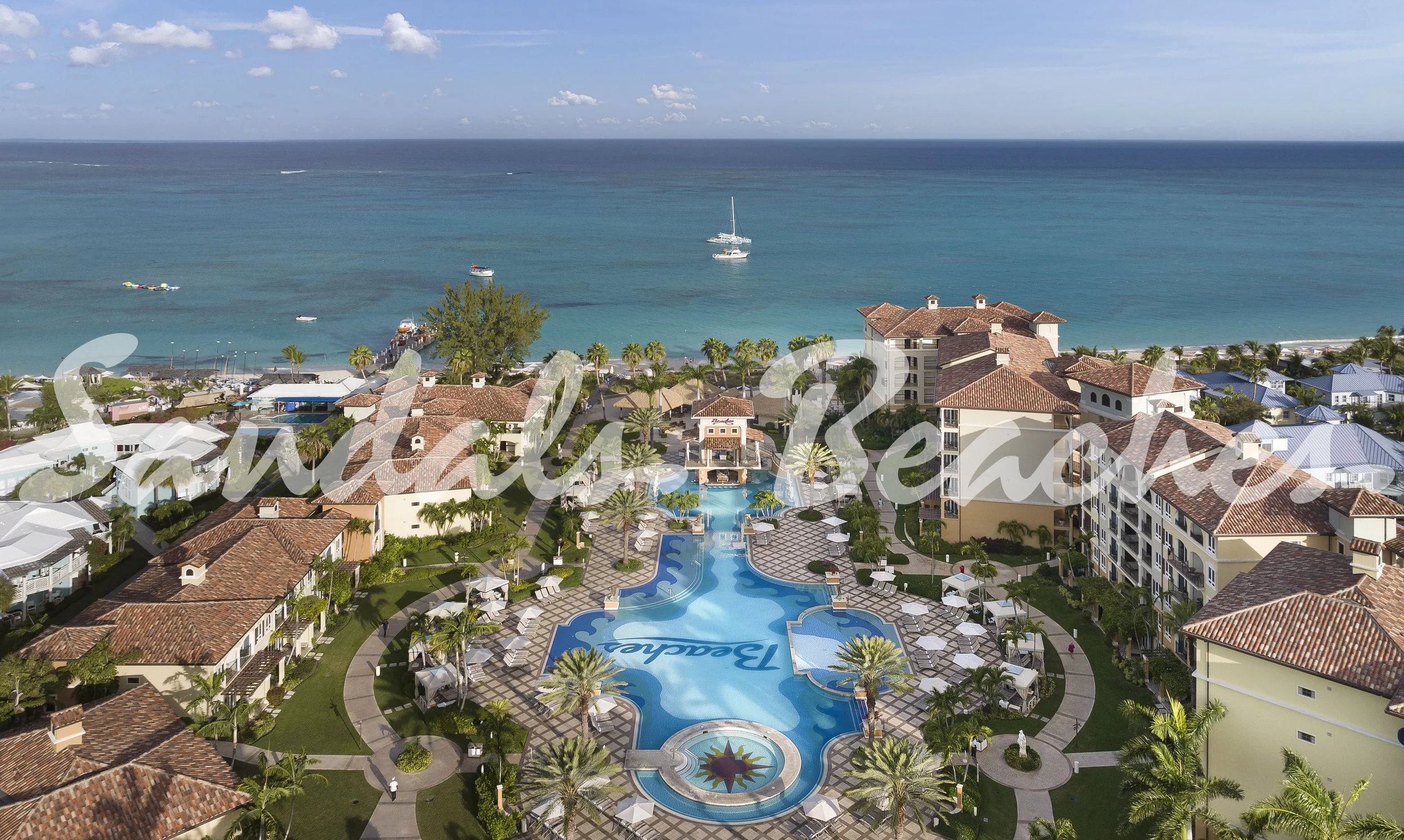Aerial view of a resort pool area with swimming pools, palm trees, lounge chairs, and umbrellas, with the ocean and boats in the background.