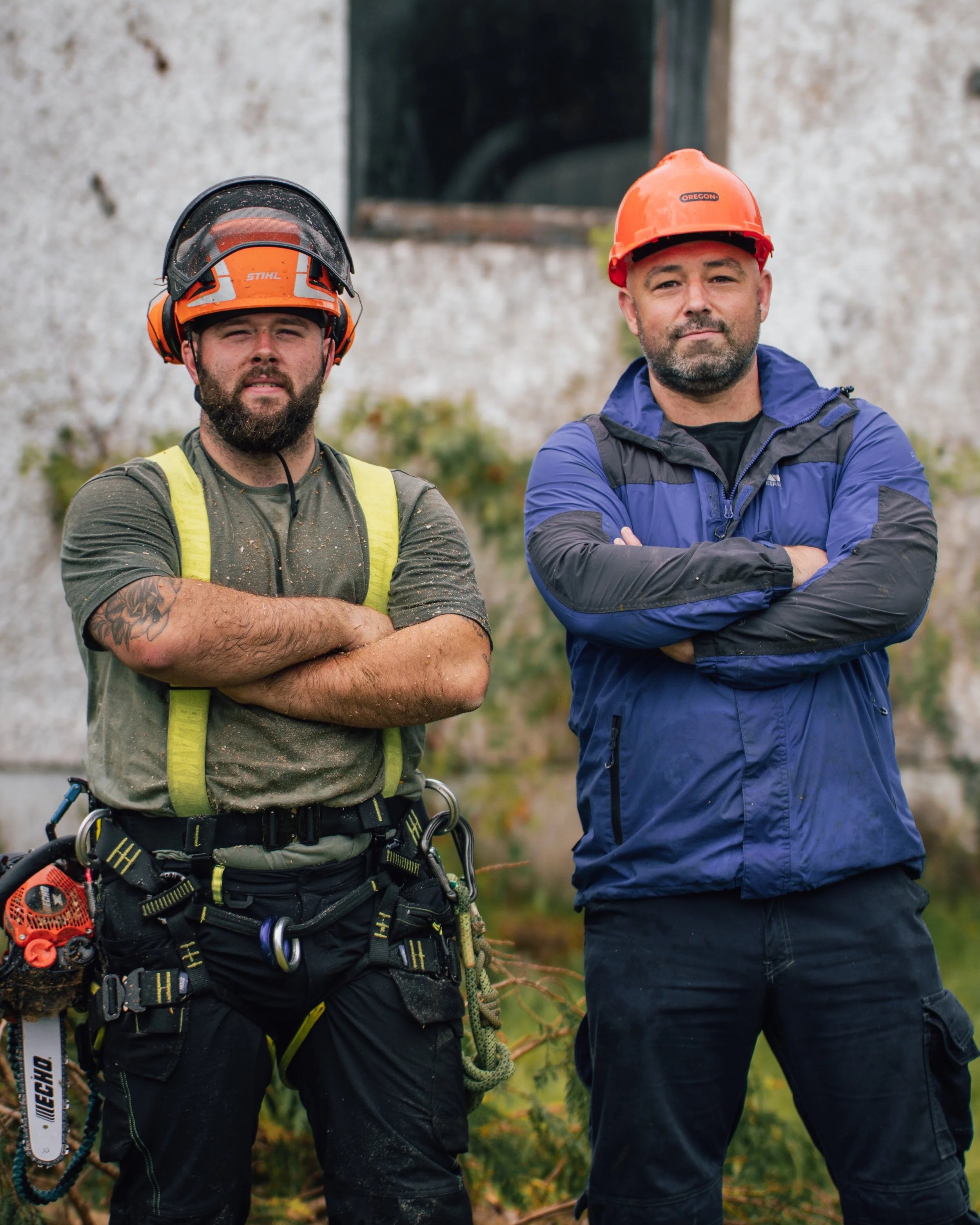 Two men standing outdoors wearing safety helmets and work clothing, with arms crossed, in front of a building with a window.