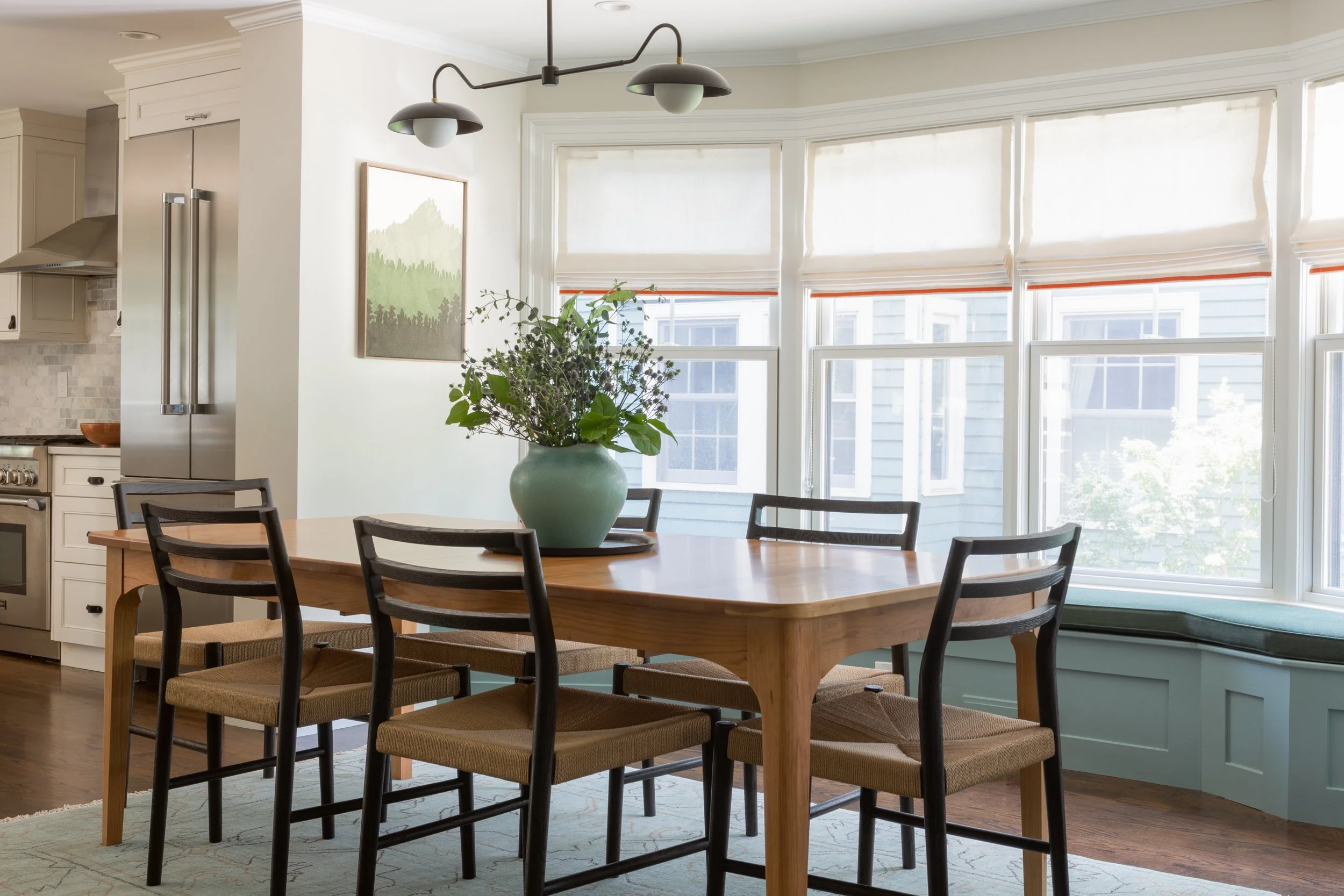 A bay window with window seat is the background for a transitional cherry dining table with black Danish-style rush-seat chairs.
