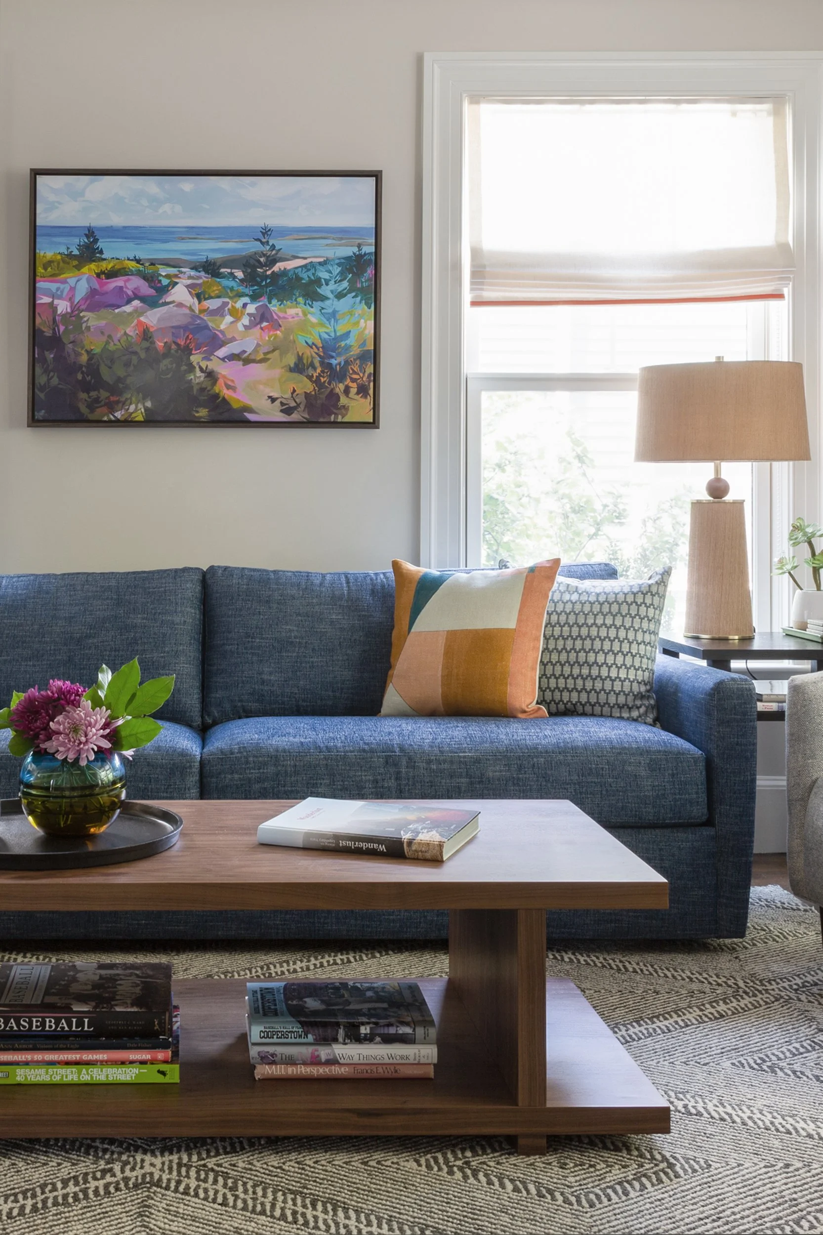 A transitional living room with a denim-blue sofa, bright Kelley Wearstler pillow, and colorful painting of Acadia National park.