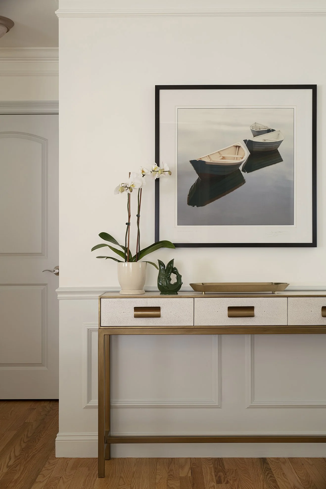 A white shagreen console sits on a wall beneath a watercolor painting of boats on a lake.