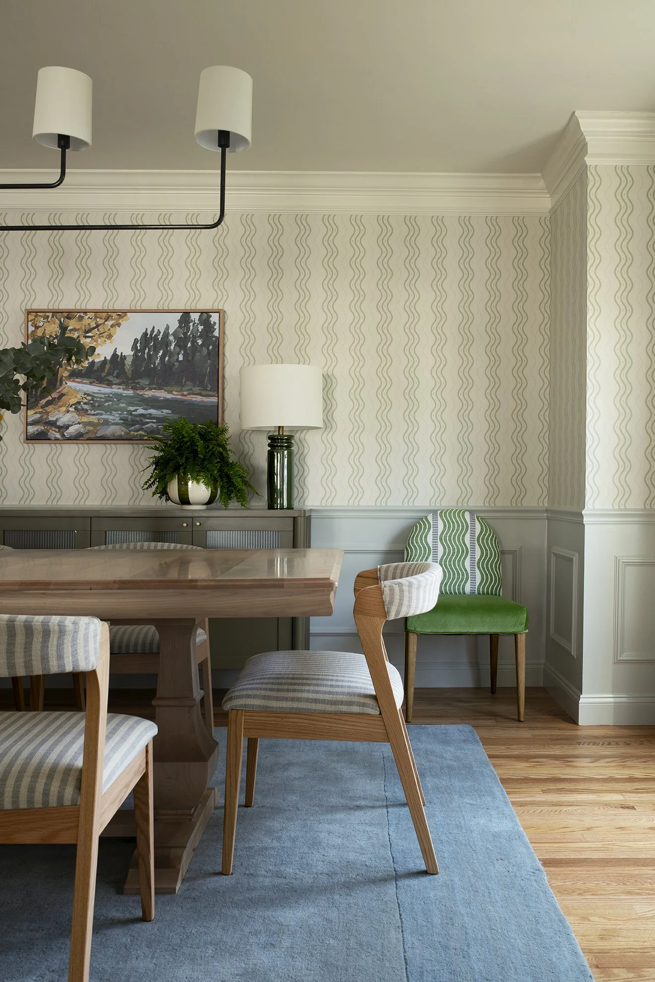 a light and airy dining room with a maple pedestal table, upholstered oak barrel back chairs, subtle striped wallpaper, and a blue rug.