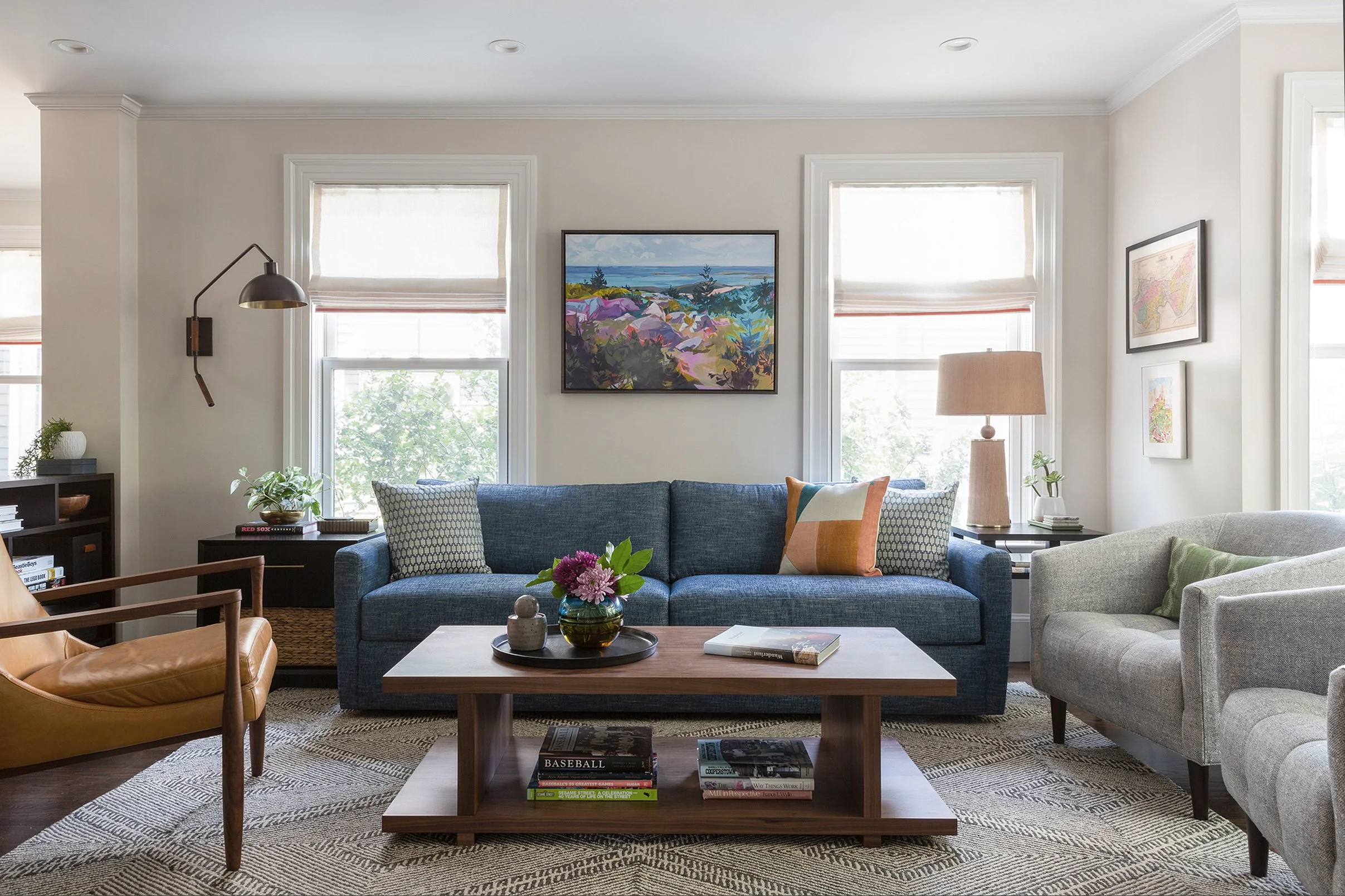 A living room with cream colored walls, a denim blue sofa, a camel leather mid-century chair, and a pair of barrel chairs. A brightly-colored painting hangs on the wall.