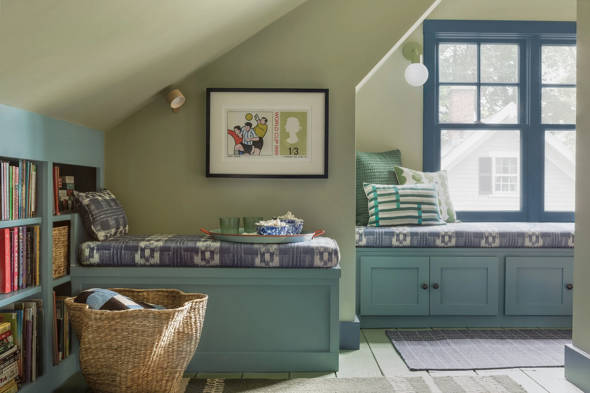 An attic reading and game room for tweens. The walls are painted Farrow & Ball Vert de Terre, with a window seat and bookshelves painted in turquoise. Cushions are upholstered in Sister Parish Mahalo, and the floor is painted green..
