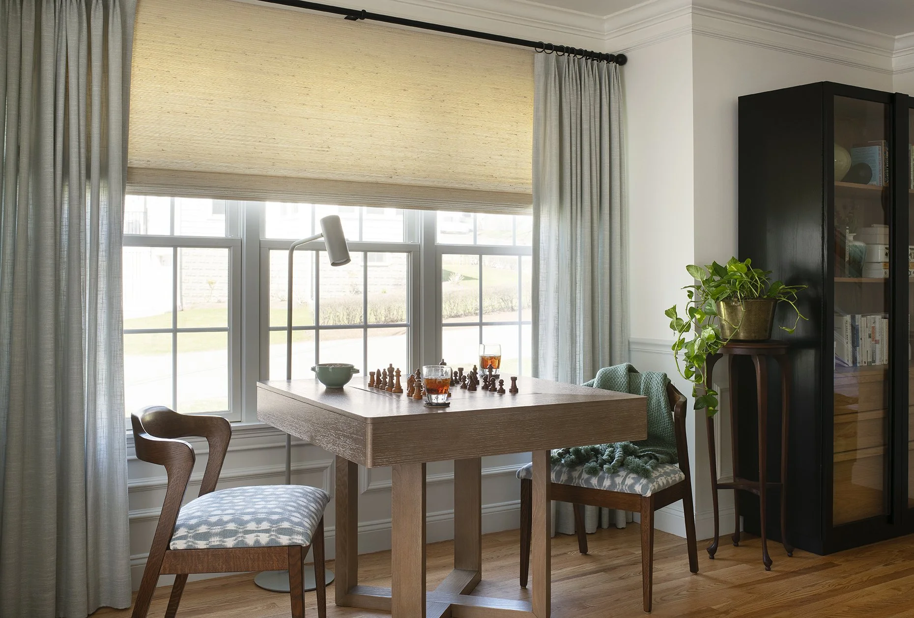 A chess table sits in front of a window. Chess pieces and drinks sit on the game table.