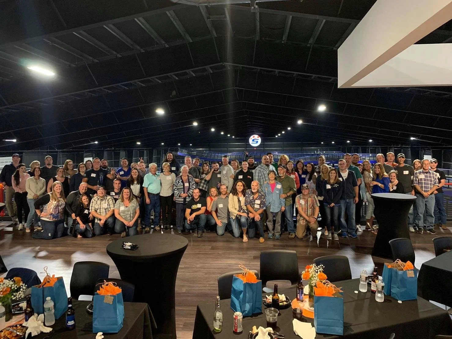 A large group of people posing for a photo in an indoor venue with a high ceiling, some tables with beverages and gift bags in the foreground.