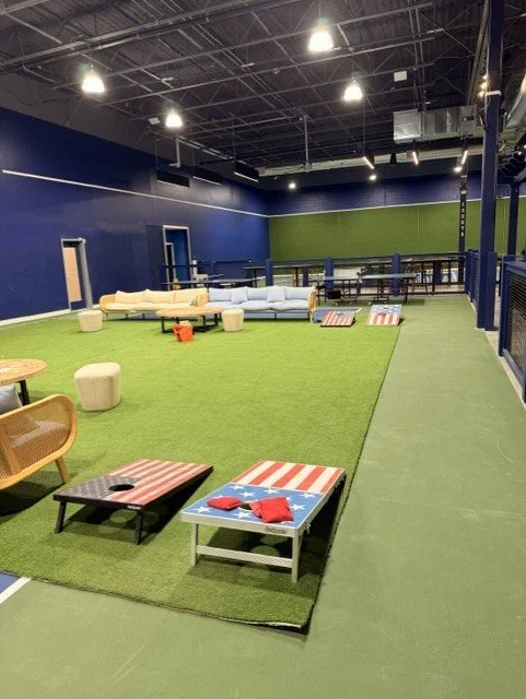 Indoor green turf area with various seating options, including small tables with red, white, and blue stripes, ottomans, and couches, in a modern recreational or lounge space.