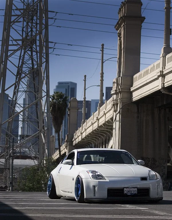 White sports car with blue wheels parked in urban setting under a bridge, with city skyline and power lines in background.