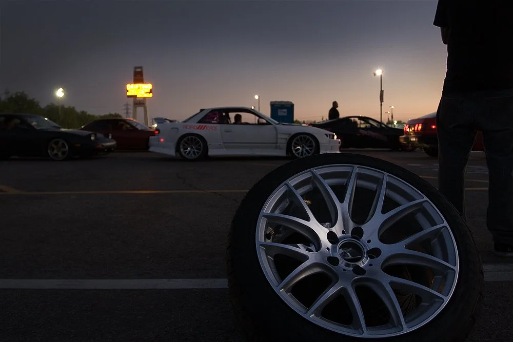 Car meet scene at dusk with sports cars and a detached silver alloy wheel in the foreground.