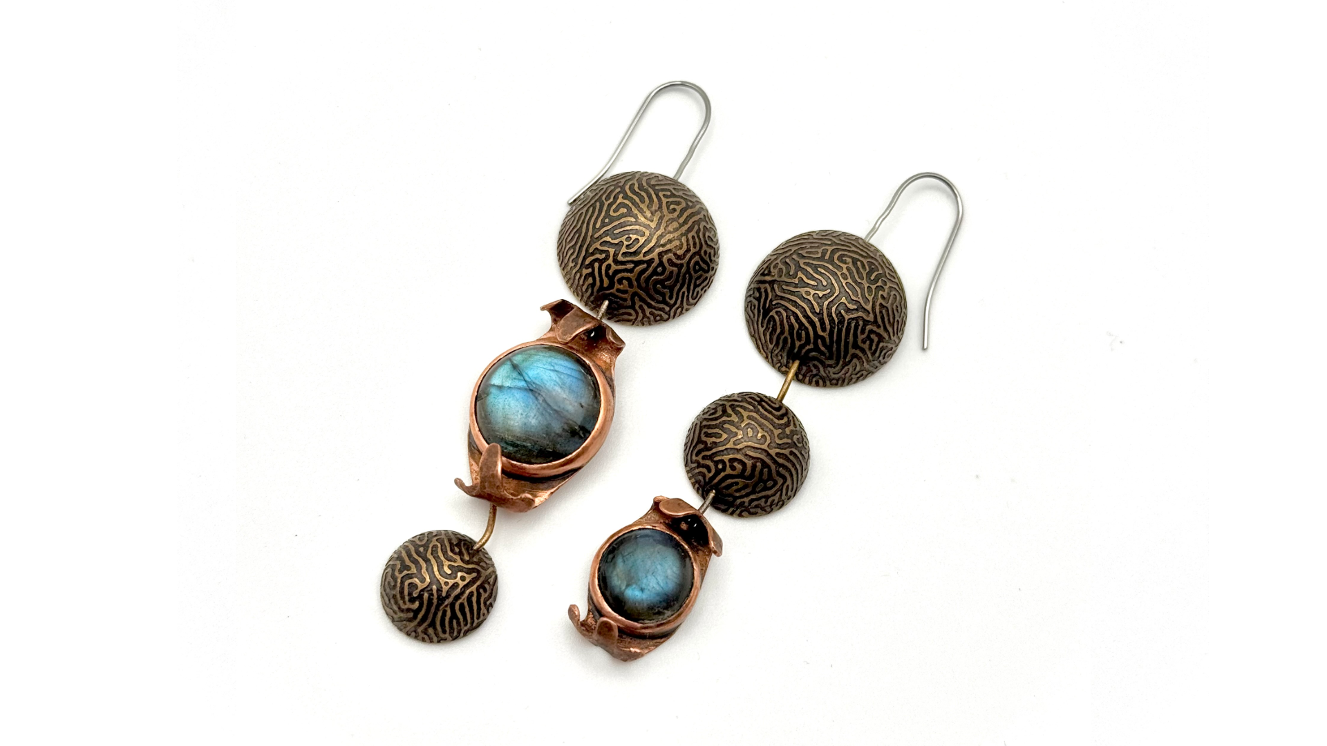 Pair of earrings with spherical patinated brass discs with intricate brain coral line patterns etched into their surface, connected to oval copper settings holding blue-green labradorite gemstones, on a white background.