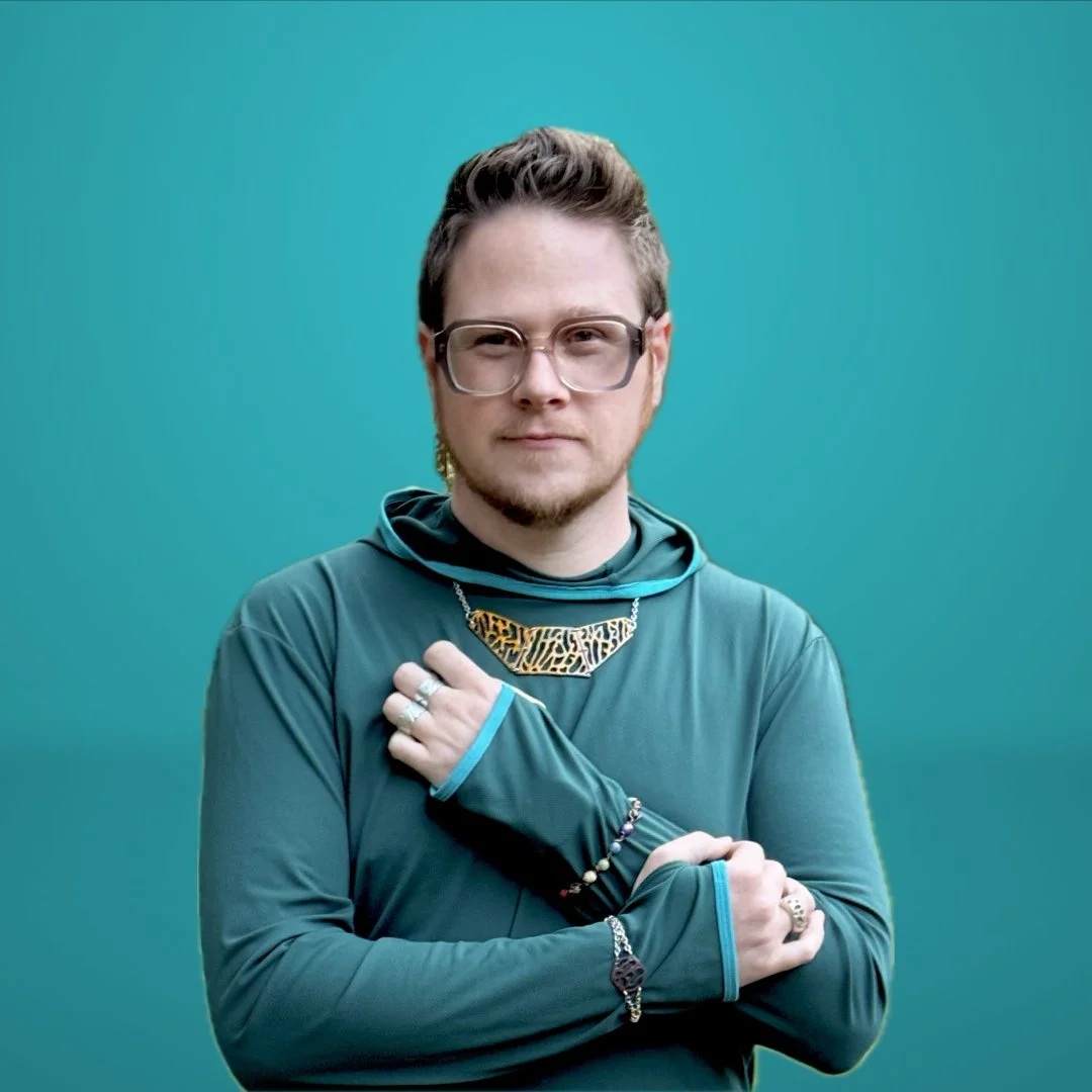 Wesley Ethyn Bowers with short hair, glasses, and a slight beard, wearing a teal long-sleeve shirt and various jewellery, standing against a teal background.