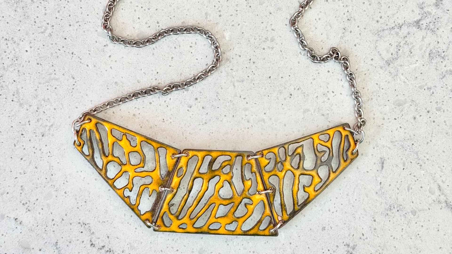 A necklace with a yellow, openwork, geometric enamel-on-copper pendant on a silver chain, placed on a light grey speckled surface.