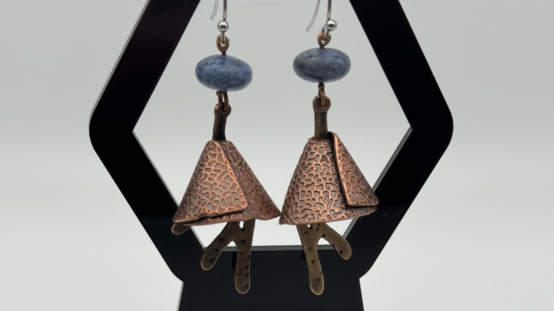 Pair of earrings with blue fossilised coral beads, copper textured conal shapes, and small dangling brass pieces, hanging in a black diamond-shaped frame.
