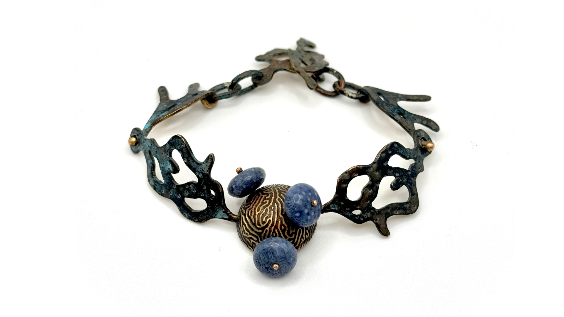 Artistic bracelet featuring a dark, abstract, coral-like metal frame with three blue spherical fossilised coral beads and a central larger oval disc with intricate brain coral patterns etched into it.