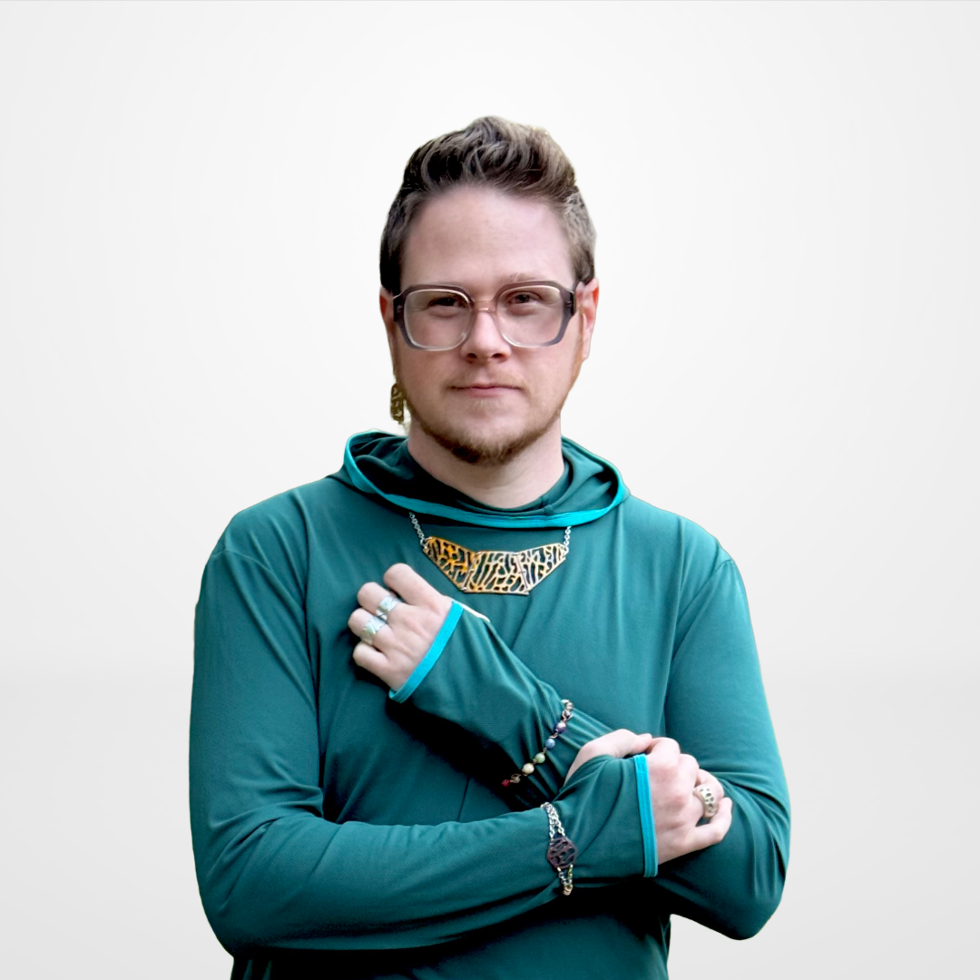 Wesley Ethyn Bowers with short hair, glasses, and a slight beard, wearing a teal long-sleeve shirt and various jewellery, standing against a light grey background.