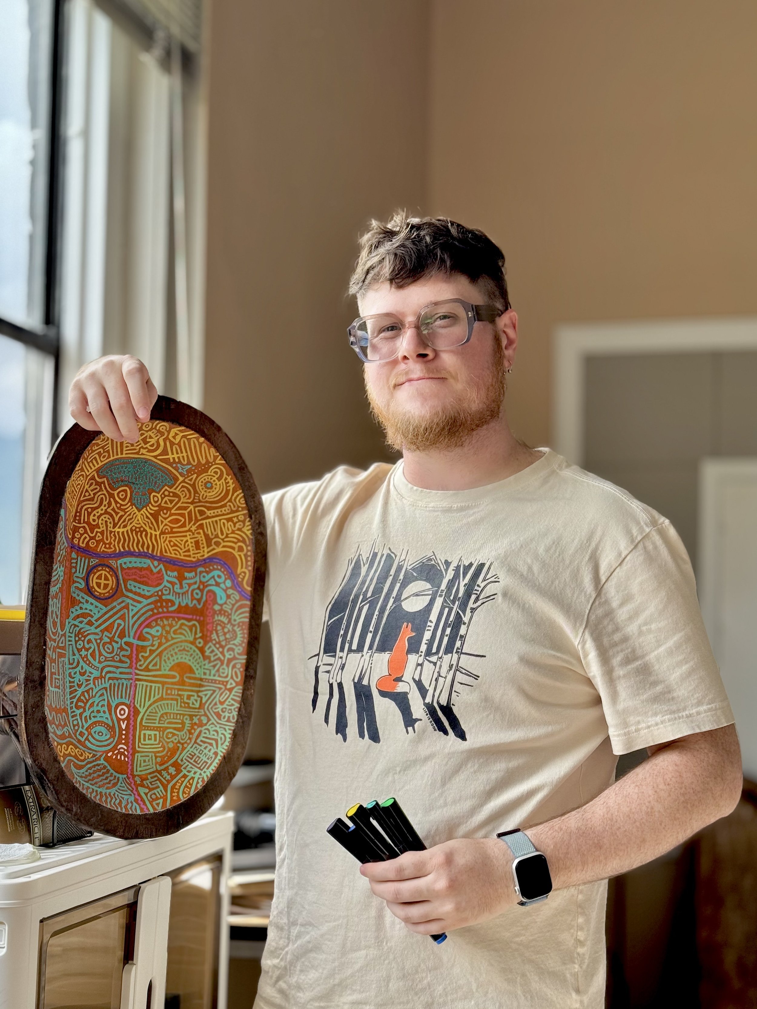 Wesley Ethyn Bowers, with glasses, a beard, and a short hairstyle, holding a colourful, intricate, oval-shaped artwork in their right hand and multiple markers in their left hand inside their studio with beige walls and a window.