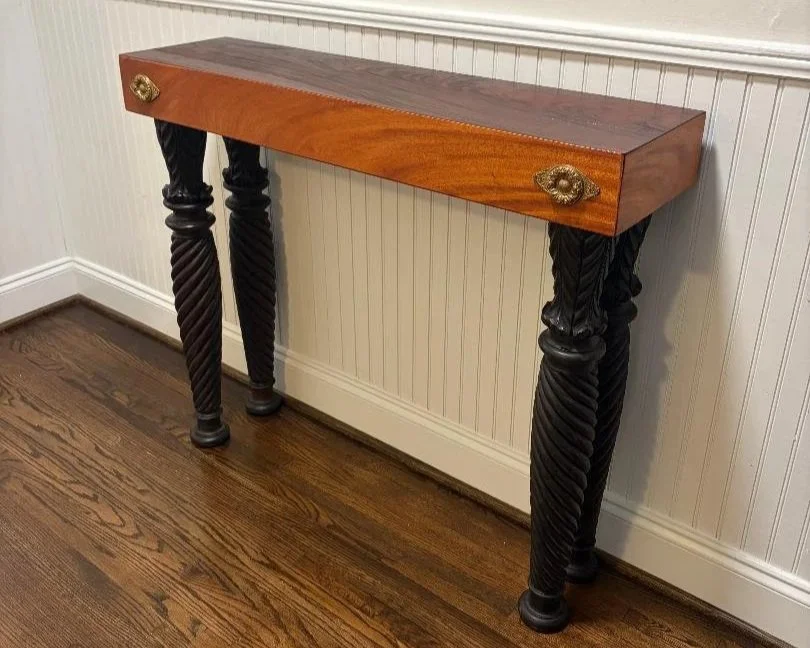 A wooden table with ornate black twisted legs and brass hardware, placed against a white wall with wooden flooring.