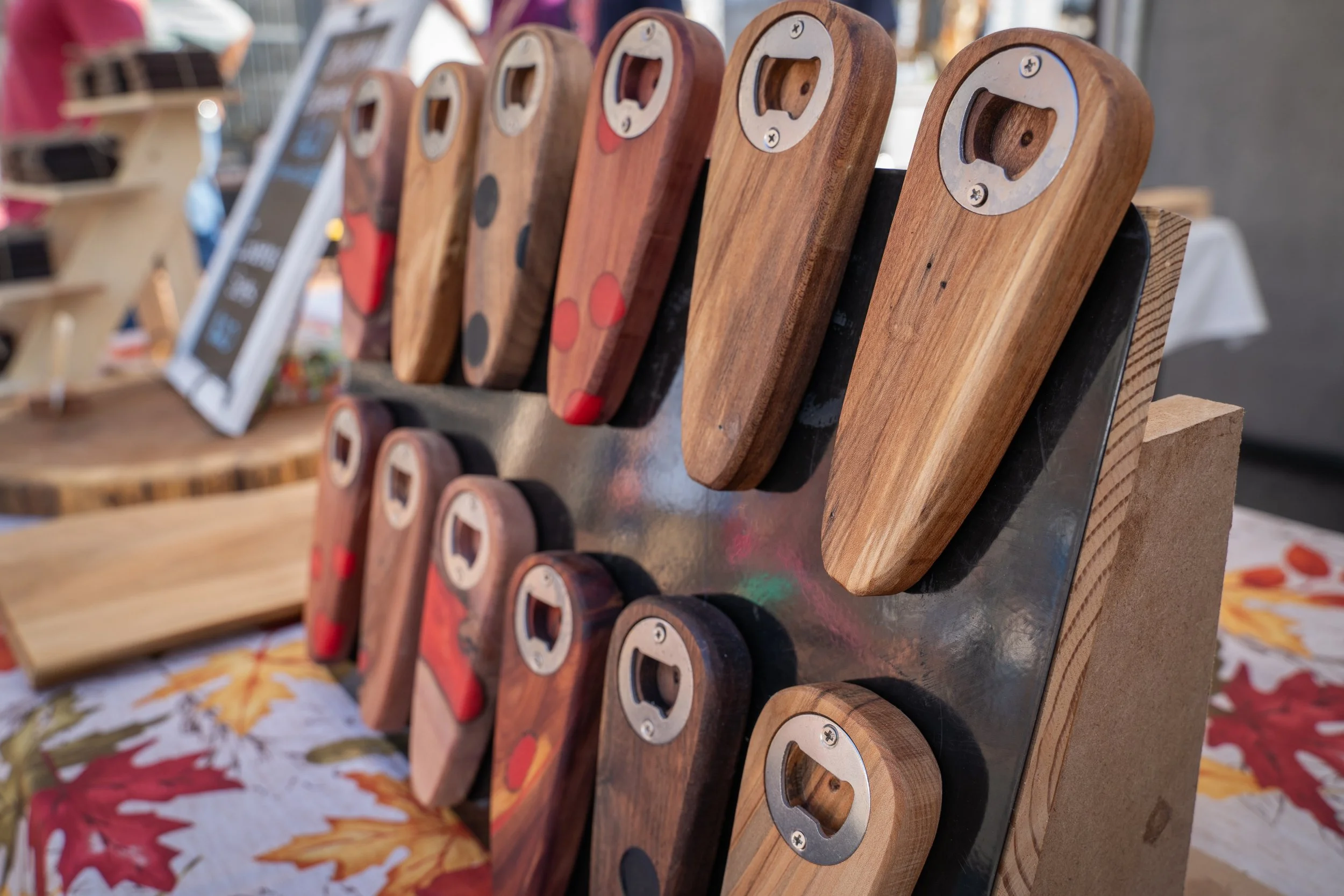 Collection of wooden bottle openers with various painted designs, displayed on a black metal stand at an outdoor market.