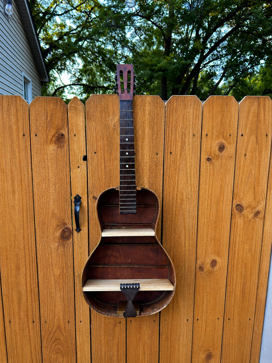 A wooden fence with an upside-down guitar attached, showing the body without strings and the neck with tuning pegs, with trees and a blue sky in the background.