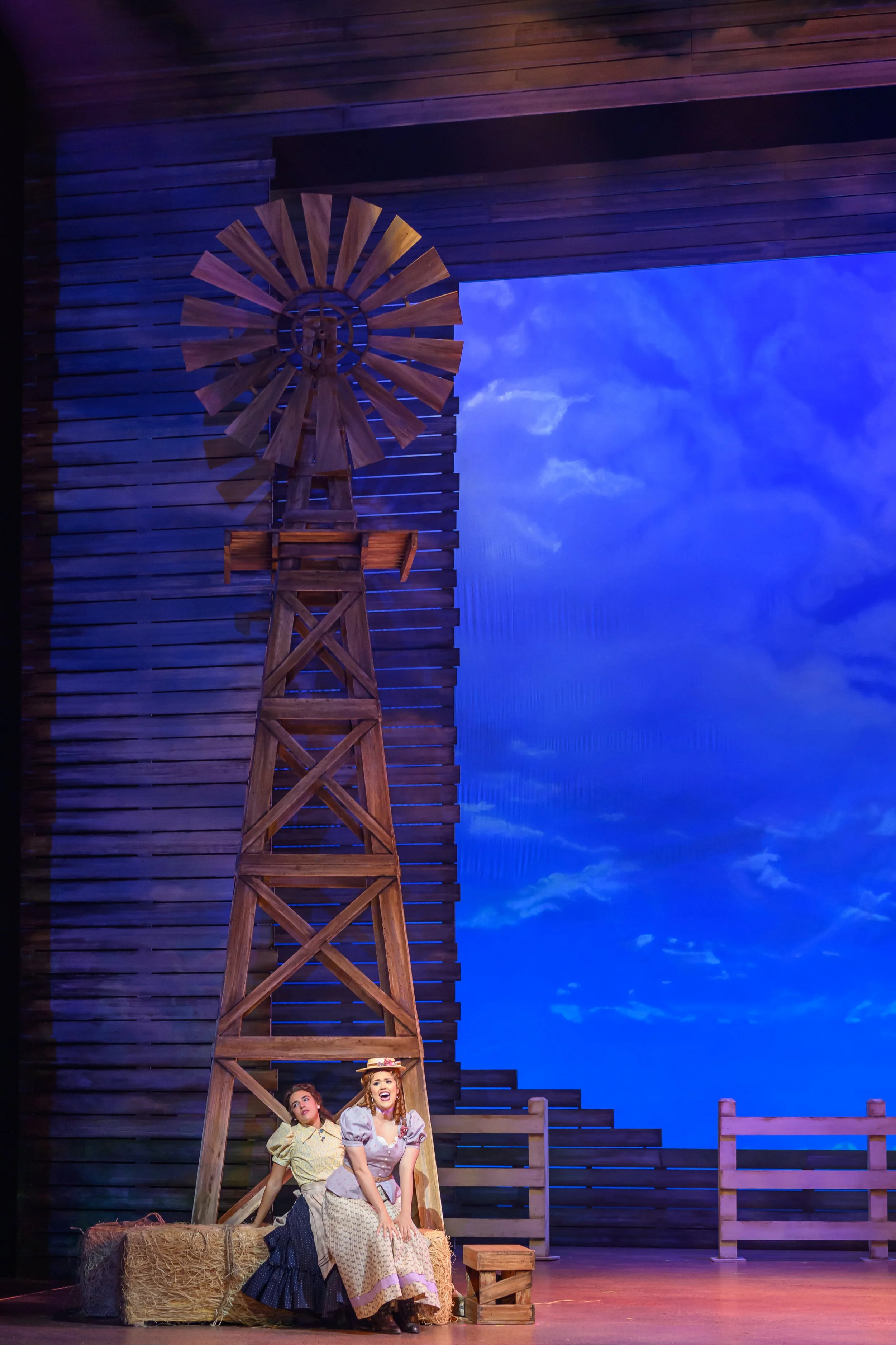 The stage scene from a musical or play set in a rural, farm-like setting featuring two women sitting on hay bales in front of a wooden windmill and barnyard fencing with a bright blue sky background.