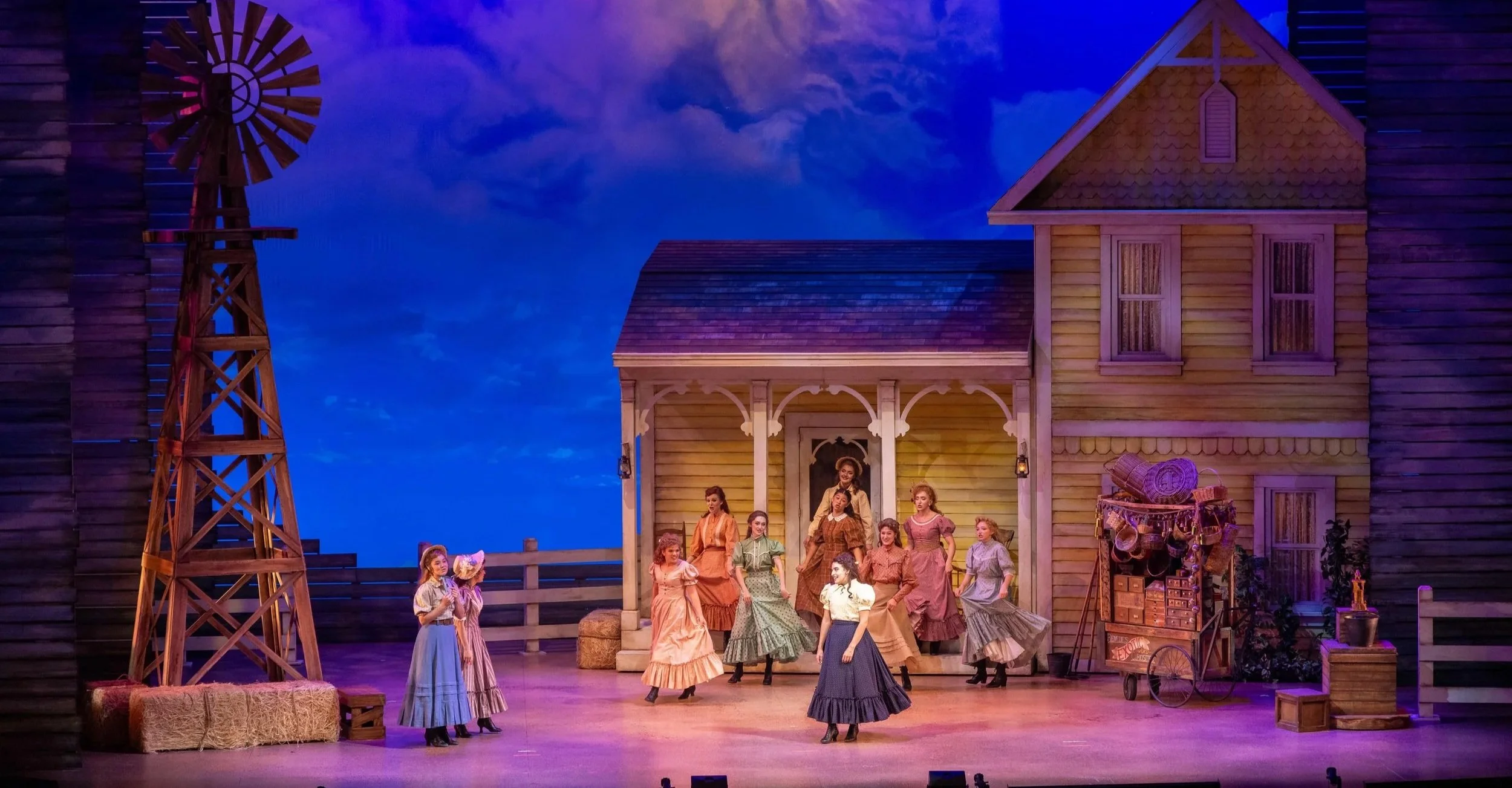 A theatrical stage with a rural farm scene, including a group of women in period costumes performing in front of a yellow wooden house with a porch, hay bales, a windmill, and farm equipment. The sky backdrop is painted to resemble dusk or early evening.