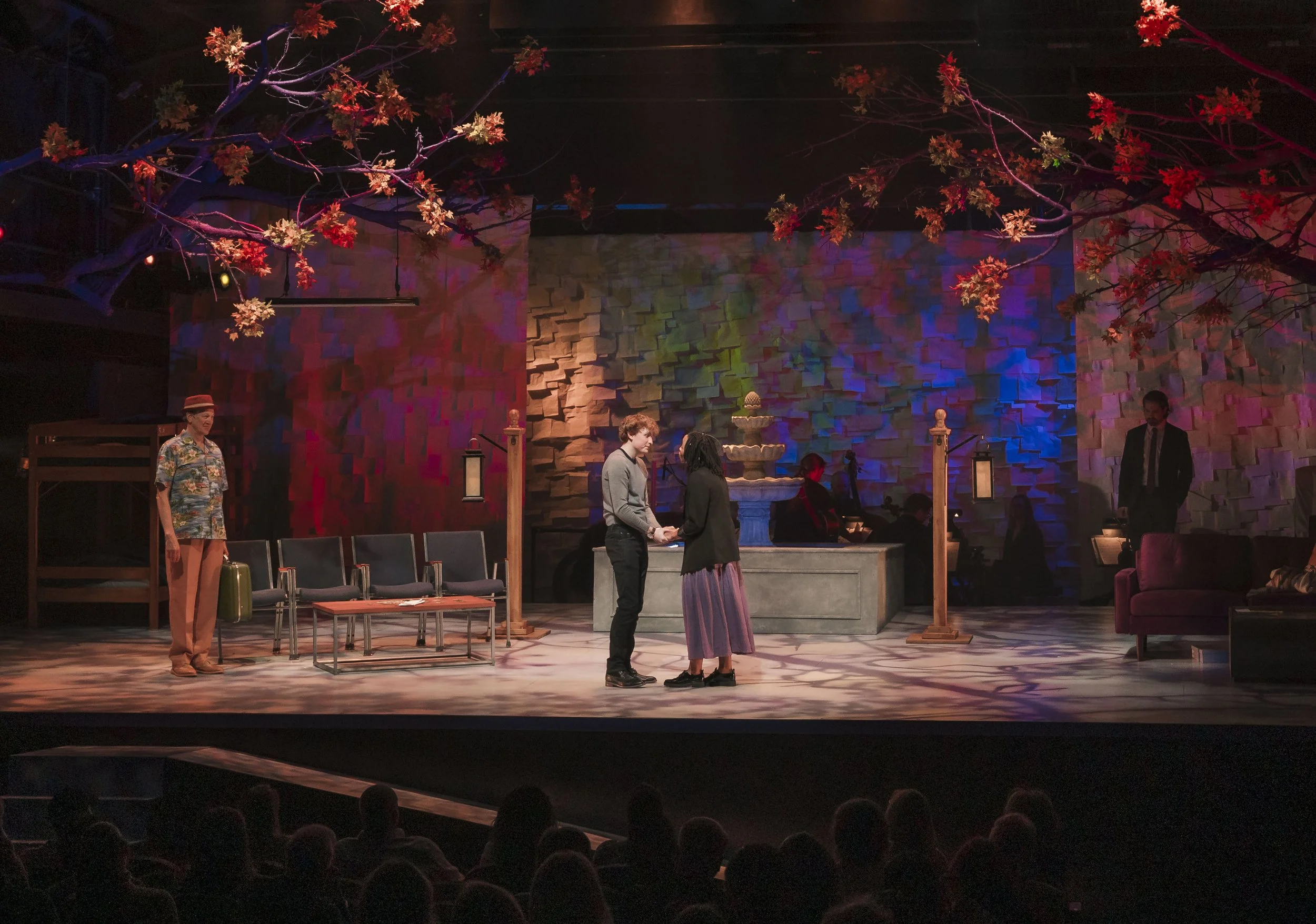 Theater stage with two people in the center holding hands, surrounded by stage scenery including trees with red leaves, a fountain, and furniture, with an audience visible at the bottom.