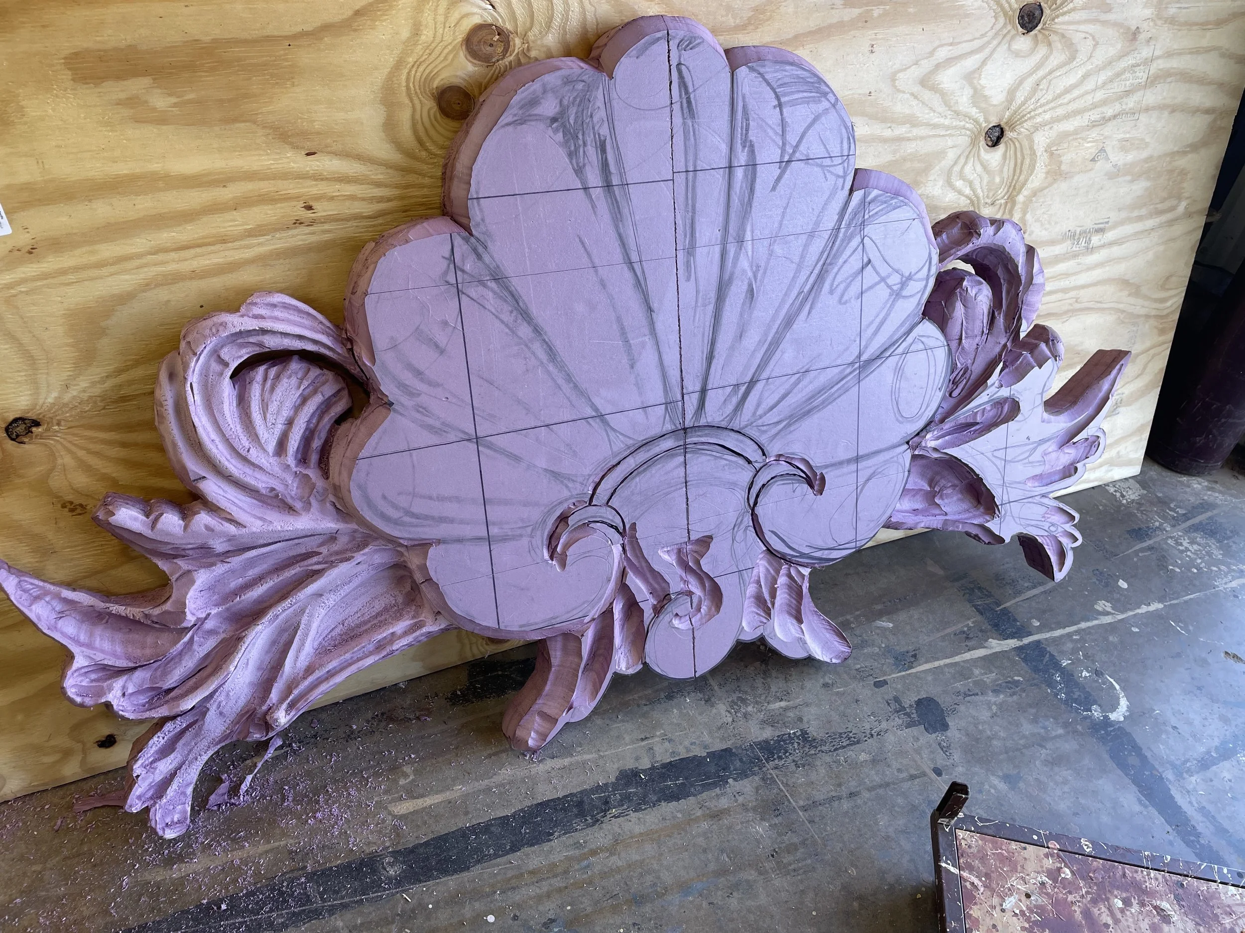 Wooden carving of a floral design in progress, with pencil and carving lines visible, mounted on a wooden background in a workshop.