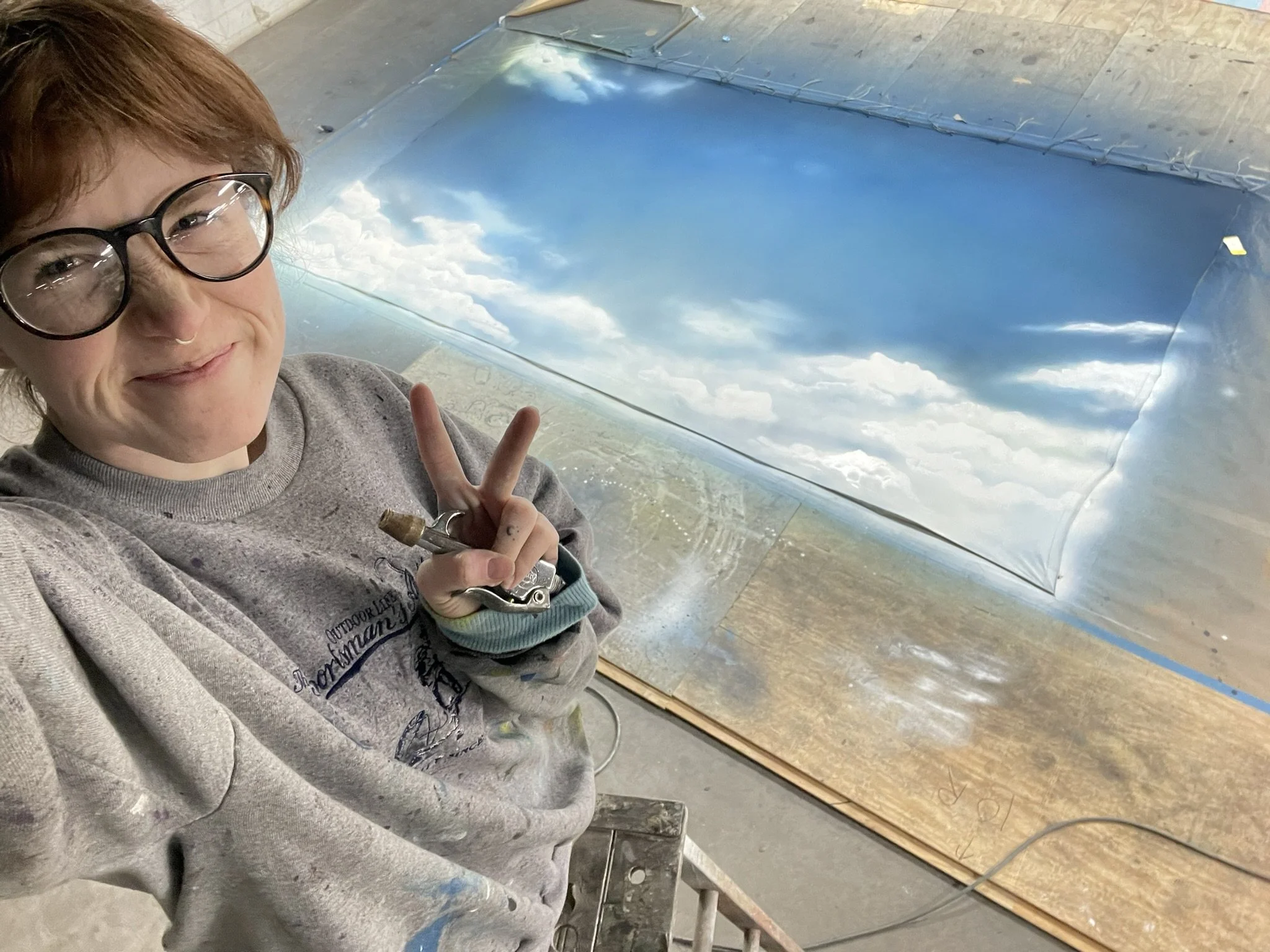 A person with glasses smiling and making a peace sign while working on an art project that involves creating a cloud scene on a large, curved, reflective surface.