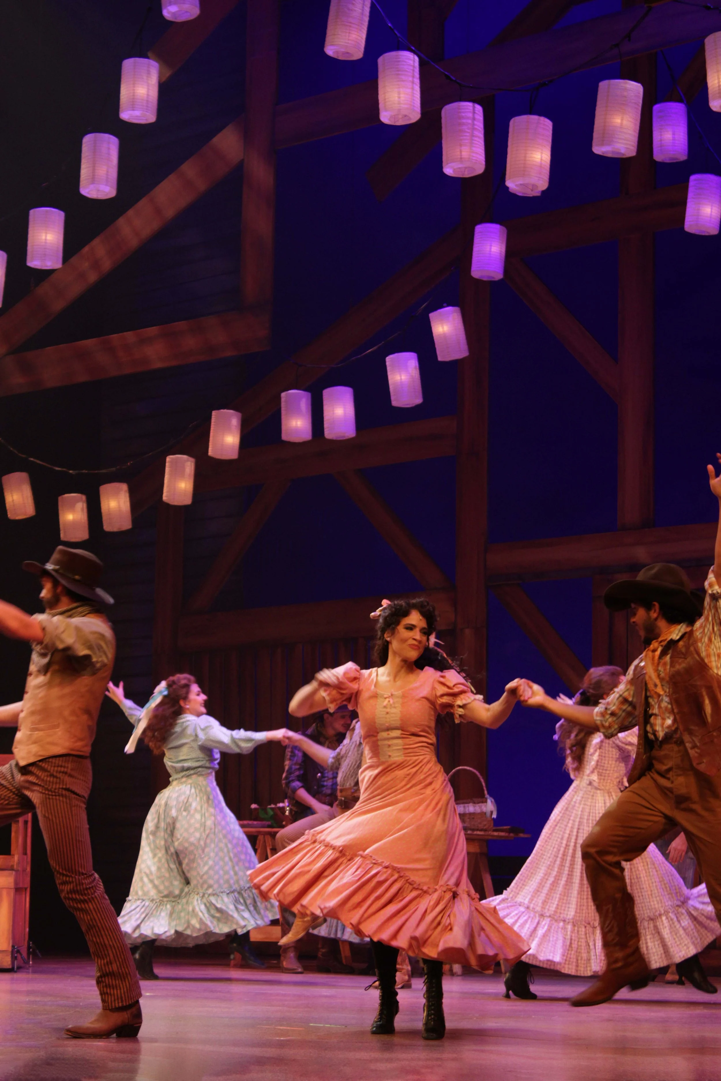 People dancing on stage with a rustic wooden barn backdrop and hanging lanterns in a theatrical performance.