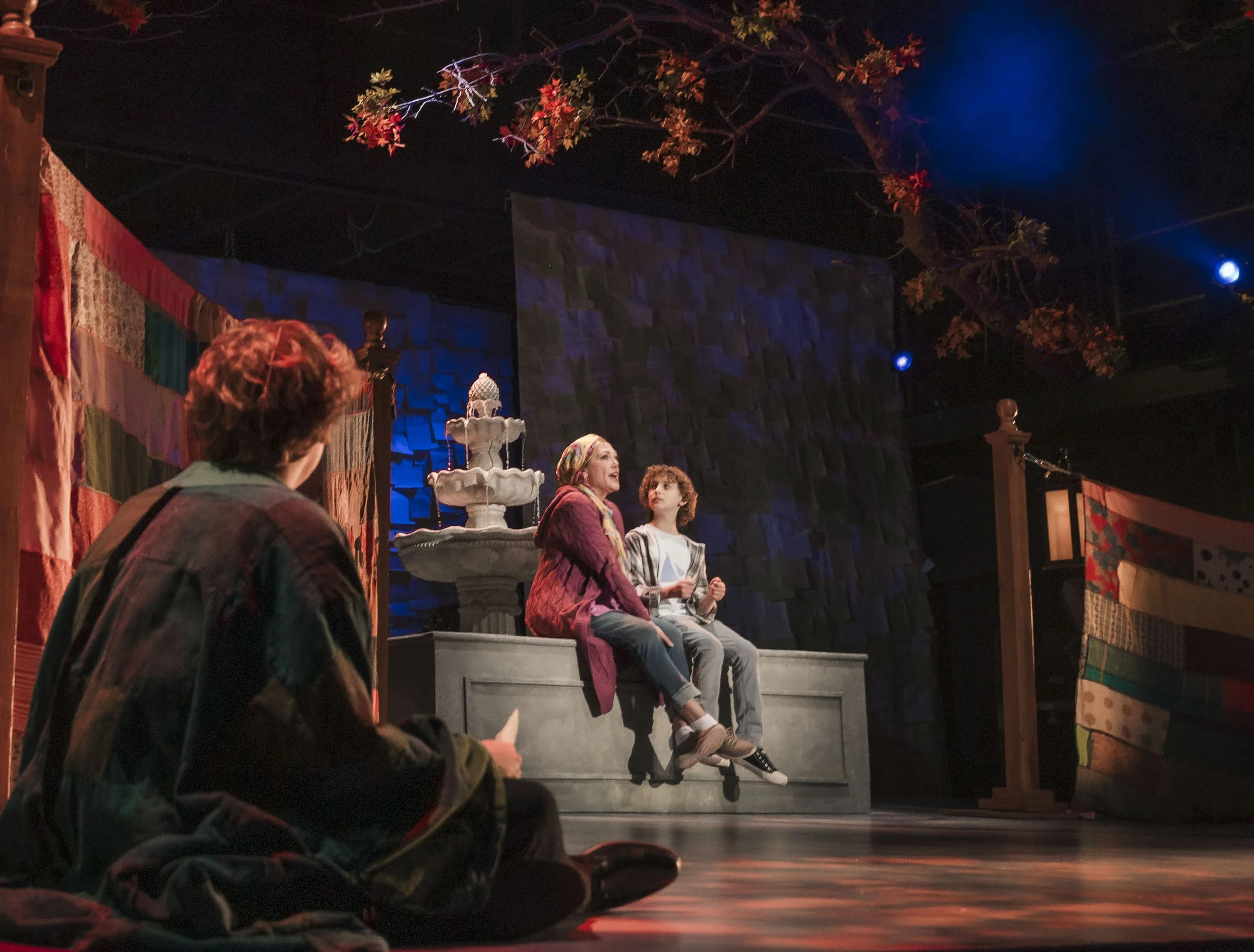 Two children sitting on a stage, with a woman speaking, decorated with colorful quilts, a fountain, and banners hanging on the sides.