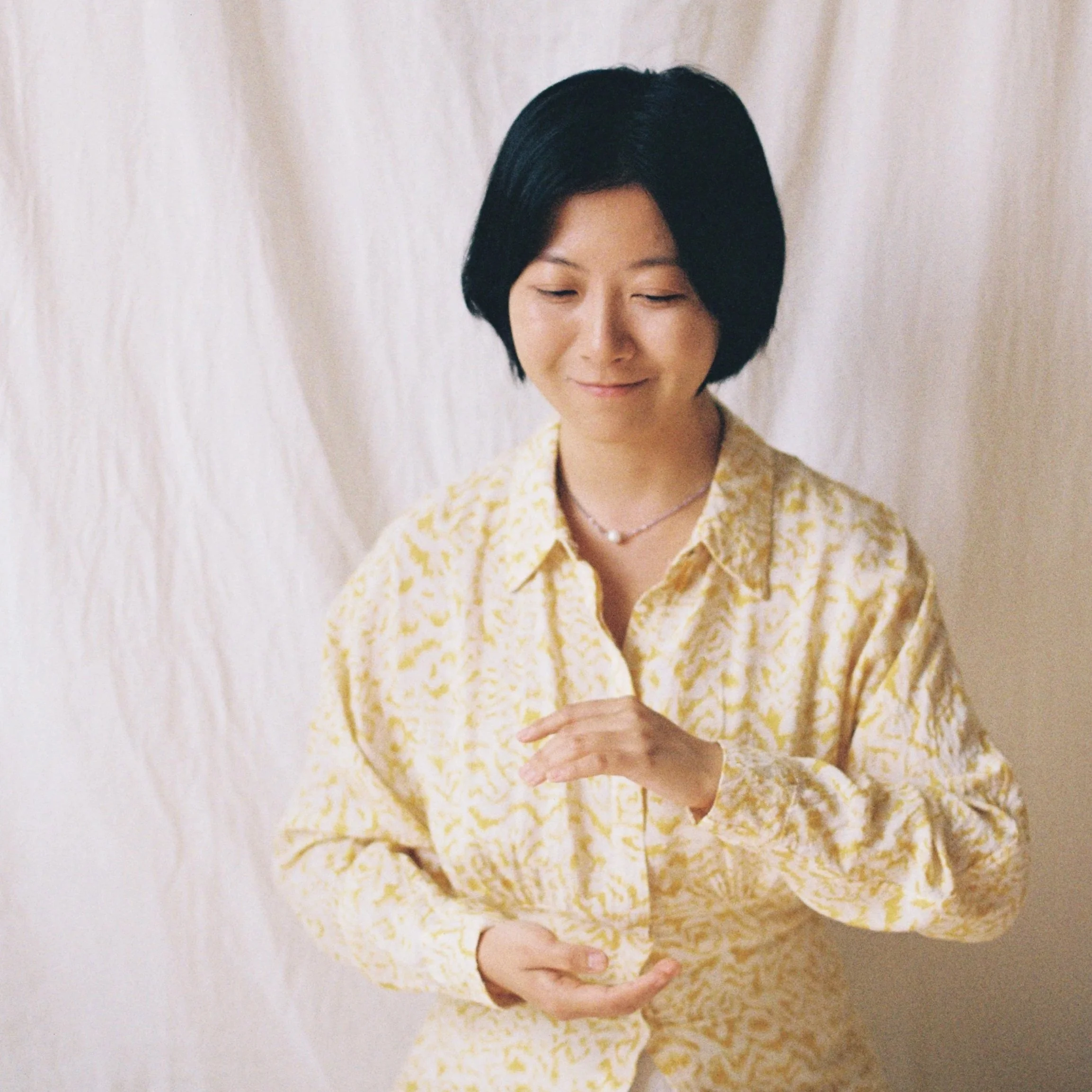 Frau mit schwarzem Bob-Haarschnitt in gelbem Kleid mit weißem Muster, die in einer Meditationspose sitzt und die Hände vor dem Bauch hält, mit geschlossener Augenpartie, vor hellem Hintergrund.