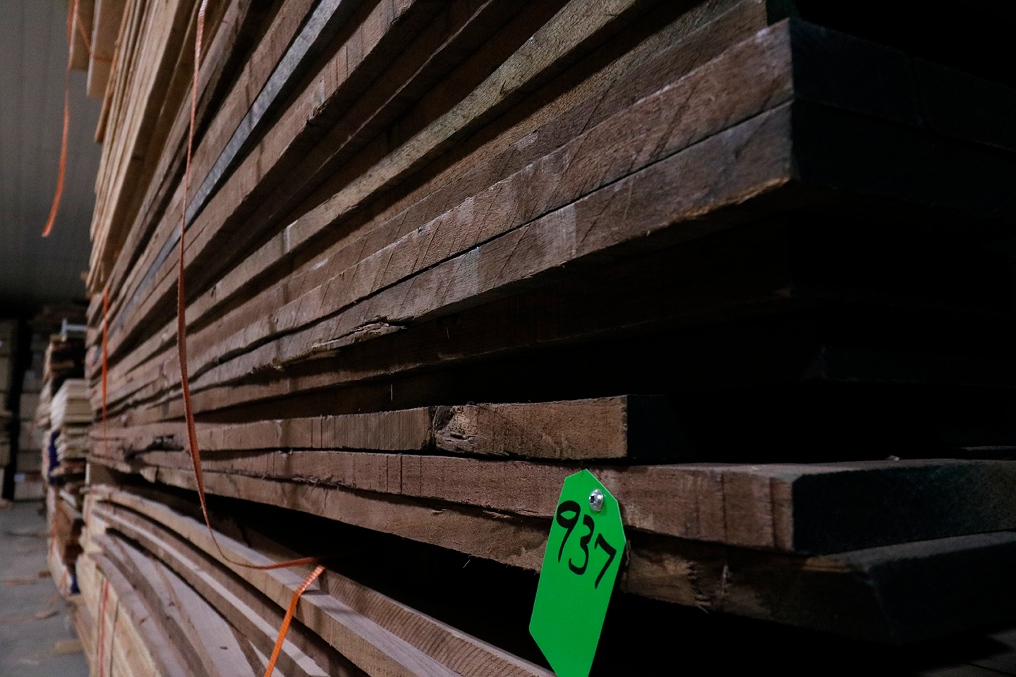Stacked wooden planks stored indoors on a warehouse shelf with a green tag numbered 937.