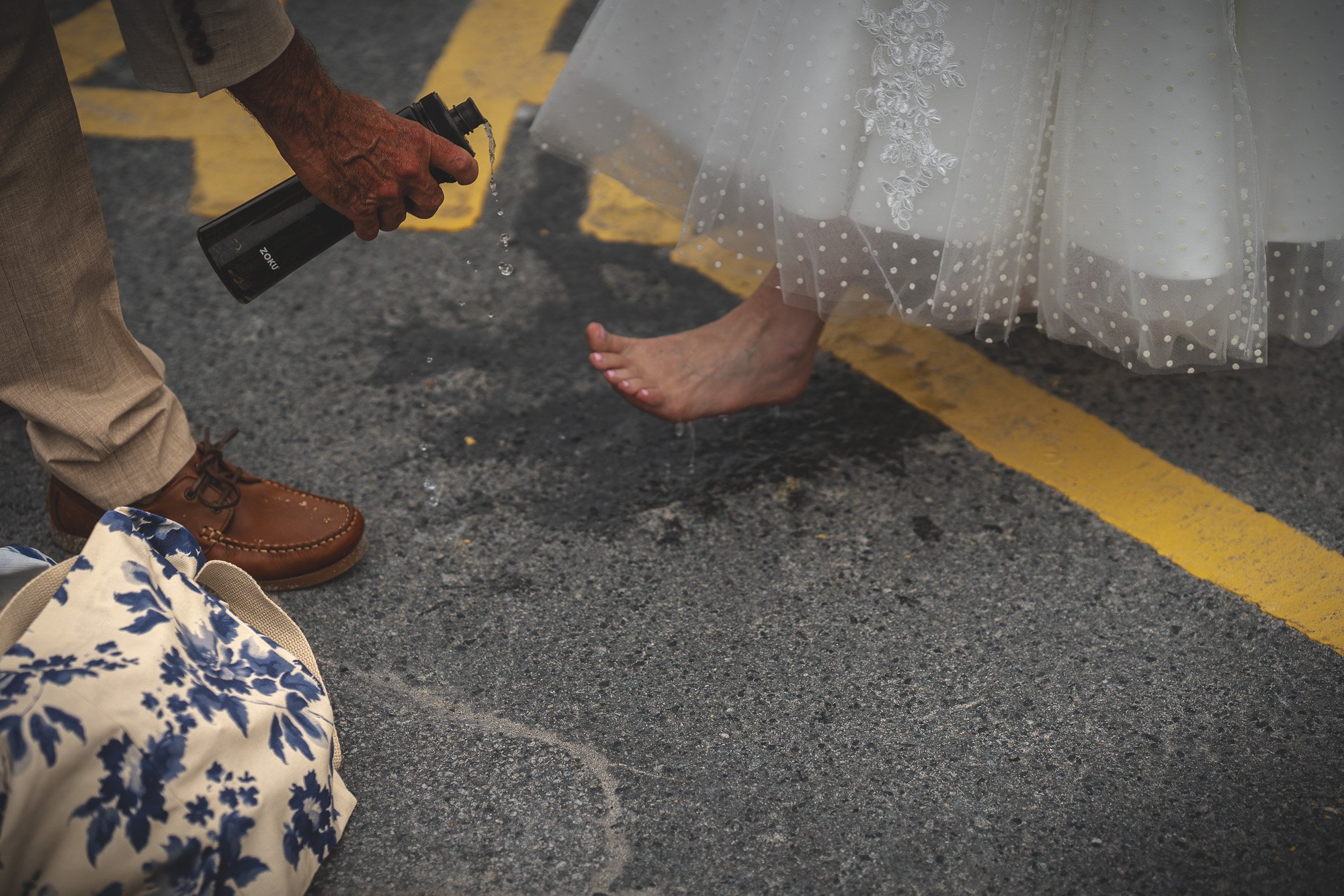 A person in formal shoes and beige pants is spraying water on a woman's barefoot foot in a wedding dress. The woman is holding an umbrella with lace detail, and a floral bag is on the ground nearby.