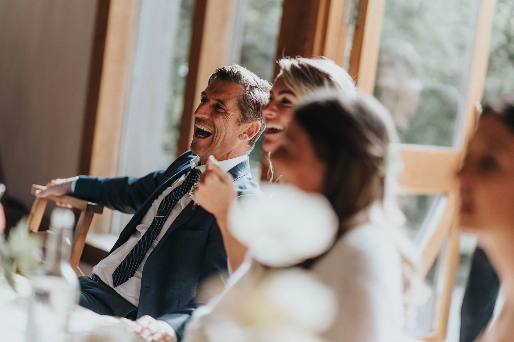 A group of people laughing and enjoying themselves at a gathering or celebration, sitting indoors near large windows with a view of trees outside.